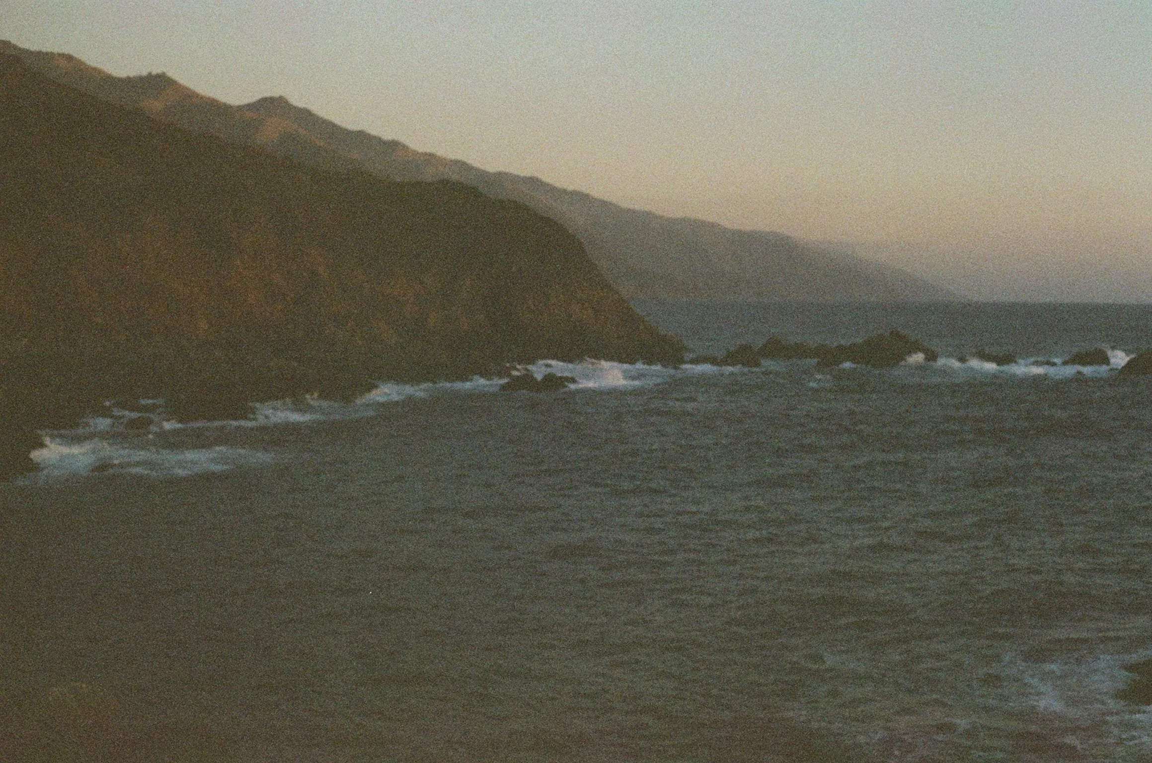 Ocean view with rugged coastline and waves, mountains in the background, dusk setting.