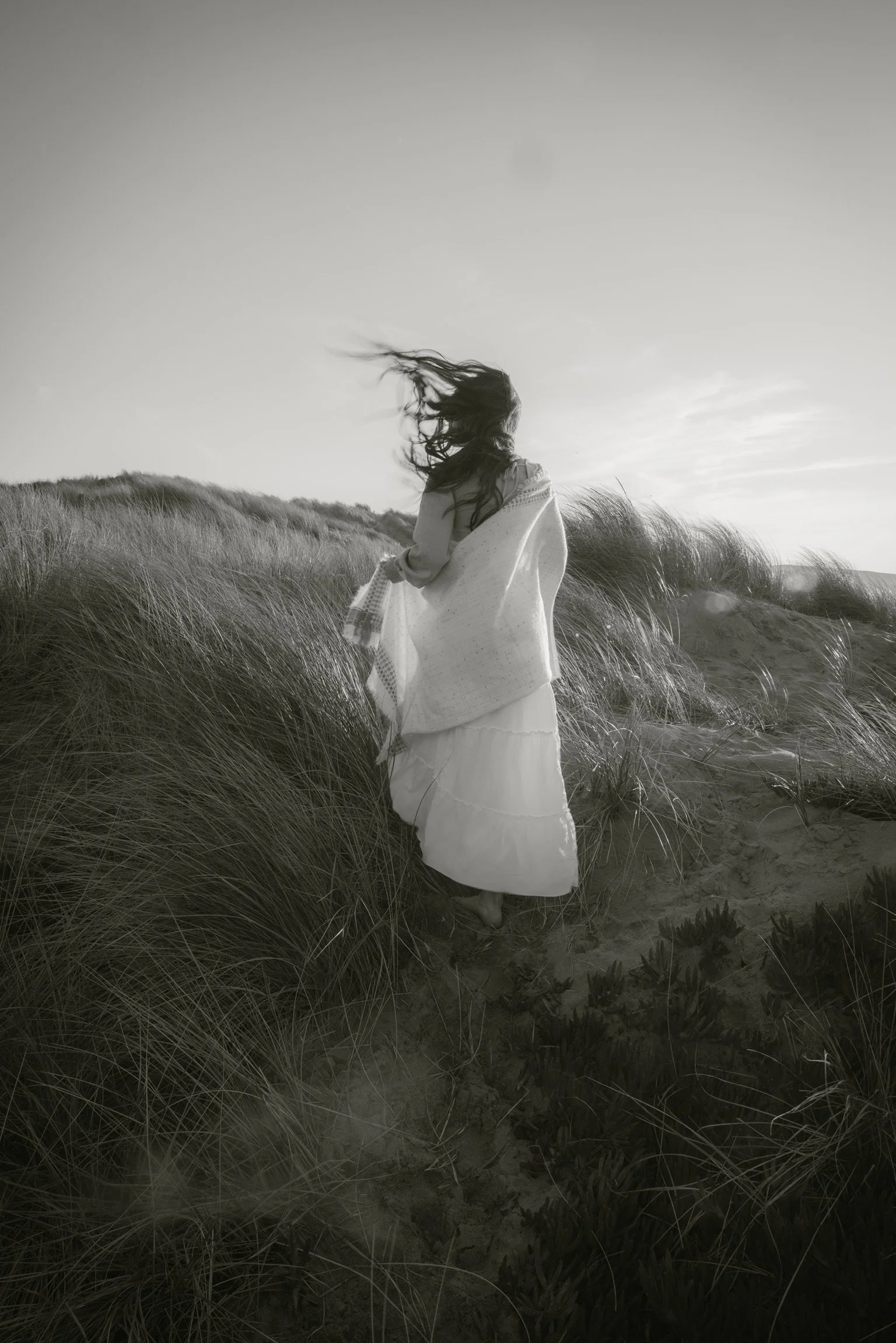 Black and white image of a woman standing on sand dunes with long grass. She is wrapped in a shawl, and her hair is blowing in the wind.