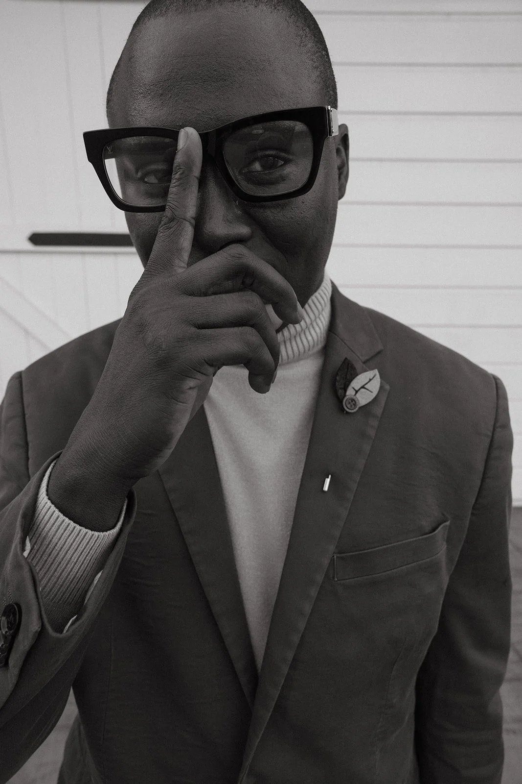 Black and white photo of a black person wearing glasses, holding their hand to their face, dressed in a suit waiting for the next food course