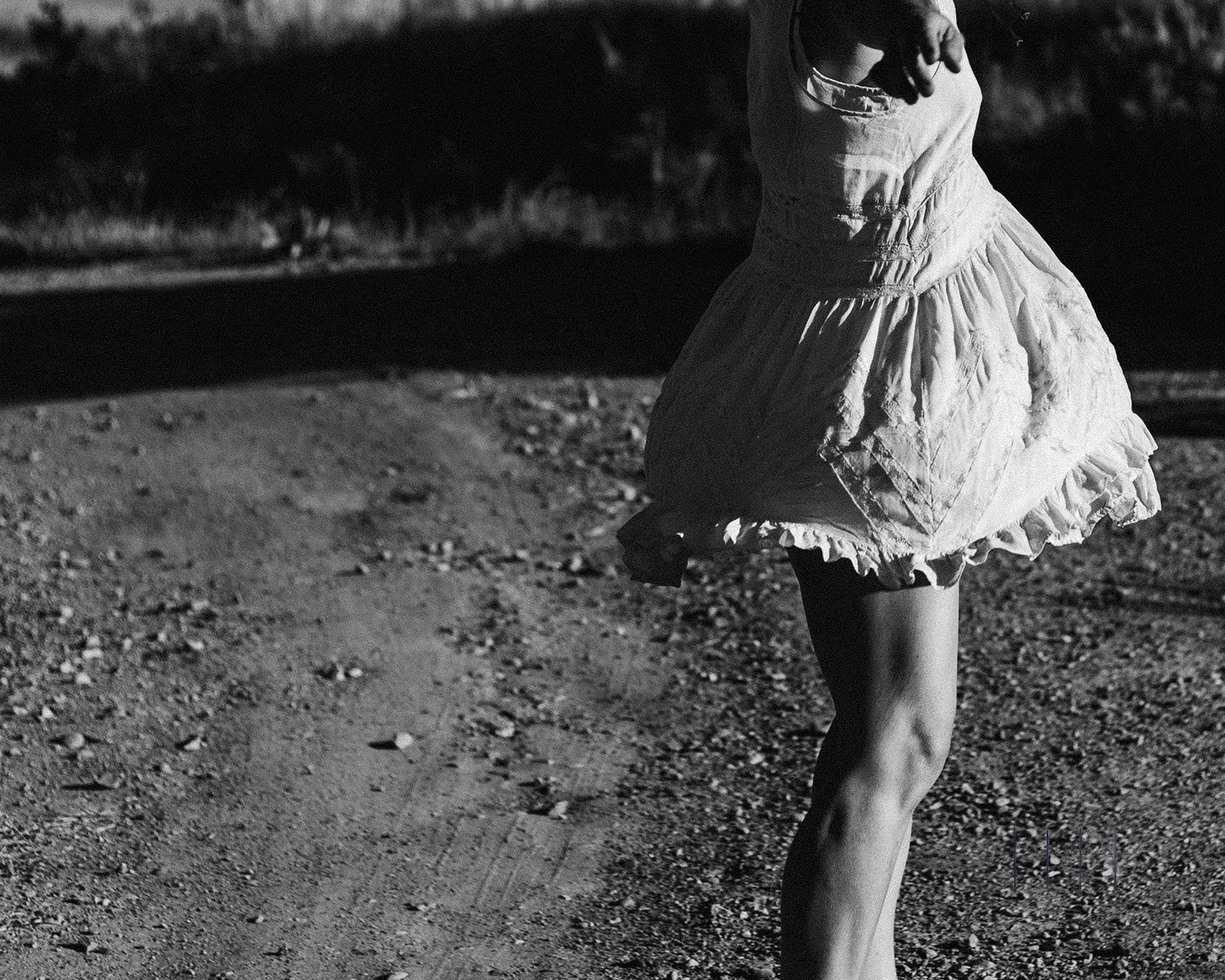Black and white photo of a person in a flowing dress dancing on a dirt path.