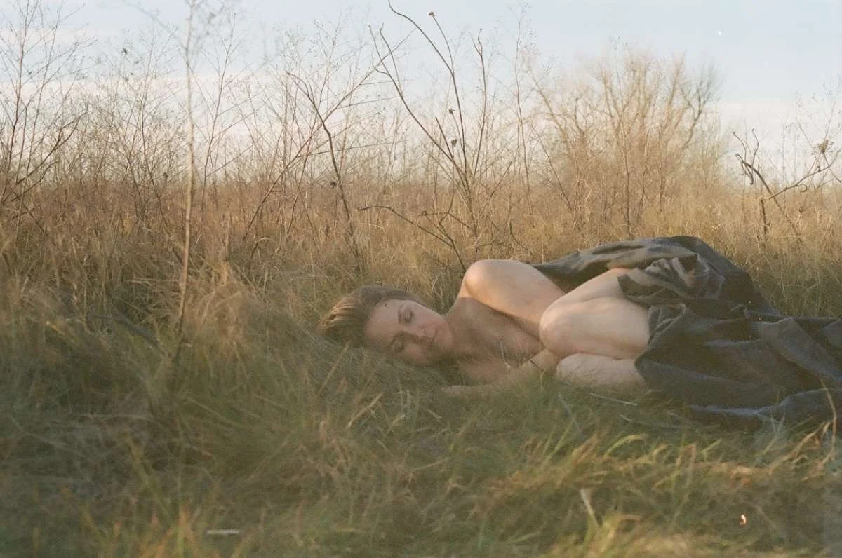 Person lying on the grass covered with a dark blanket in a field with dry vegetation.