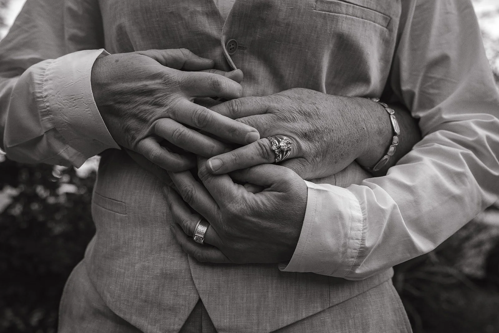 Two people wearing formal clothing embrace, showing hands with rings and a bracelet.