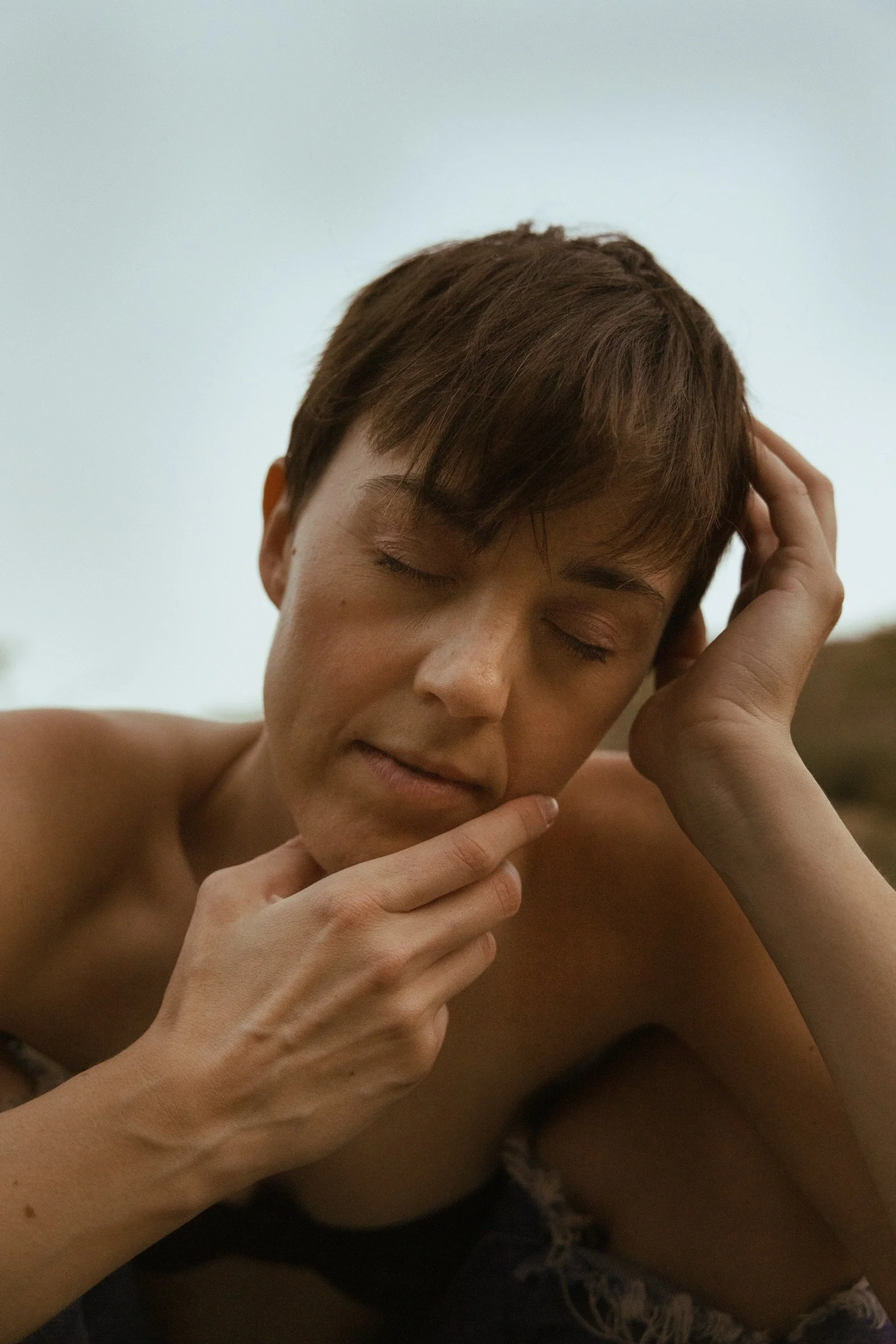 Person with short hair resting their head in one hand and touching their chin with the other, eyes closed, against a soft-focus background.