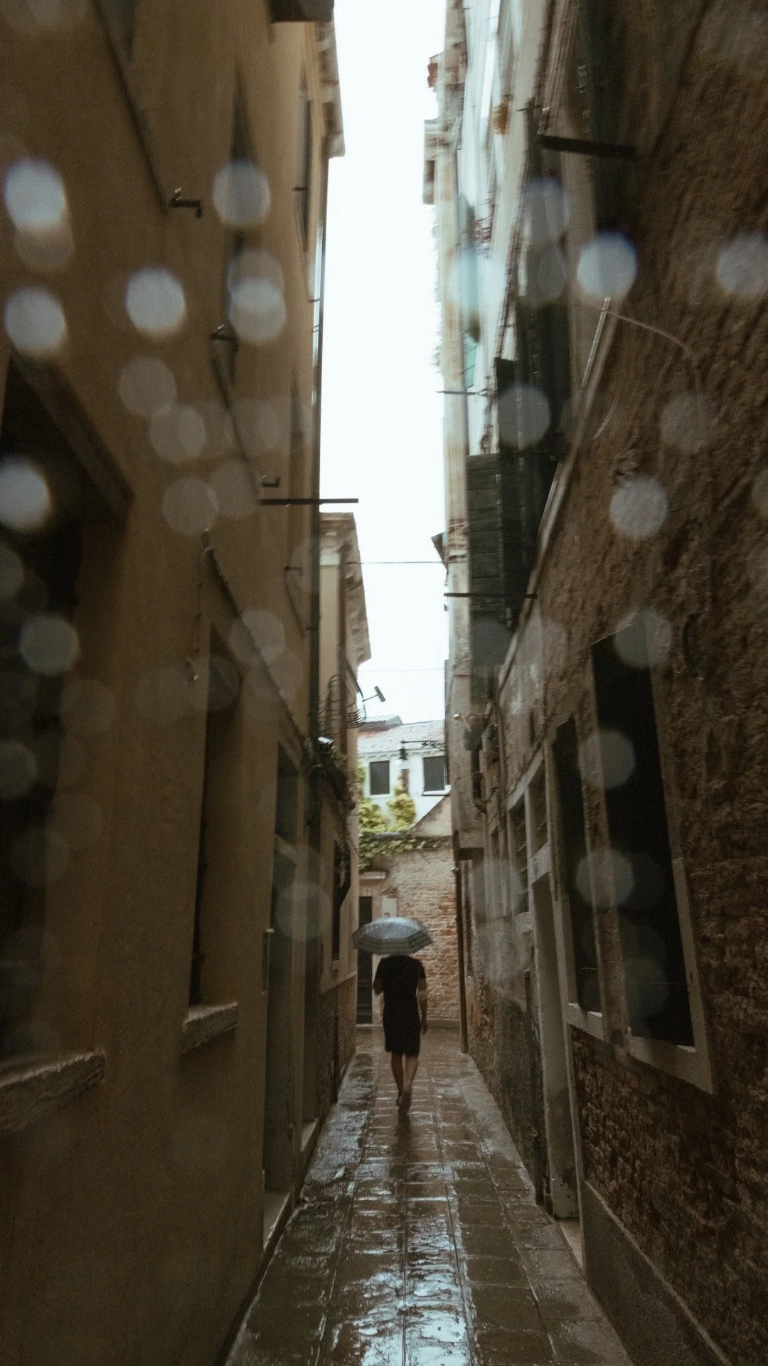 Person walking with an umbrella in a narrow alleyway with wet cobblestone pavement, blurred raindrops visible.
