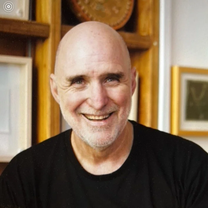 A smiling, bald man with a white beard wearing a black shirt, in front of wooden shelves with framed pictures and decorative items.