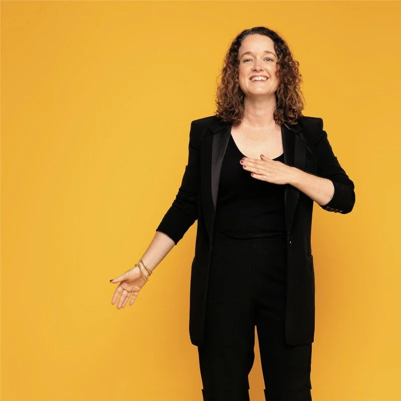 Woman with curly hair, wearing a black blazer and pants, smiling, with her right hand on her chest and her left arm extended, against a yellow background.