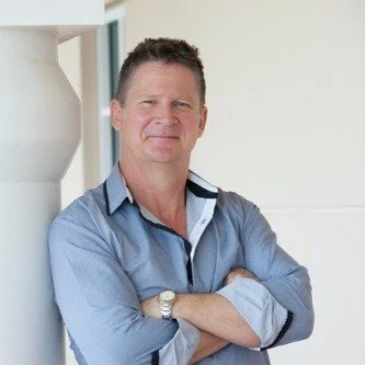 Man in a light blue shirt standing with arms crossed, leaning against a wall in an indoor setting.