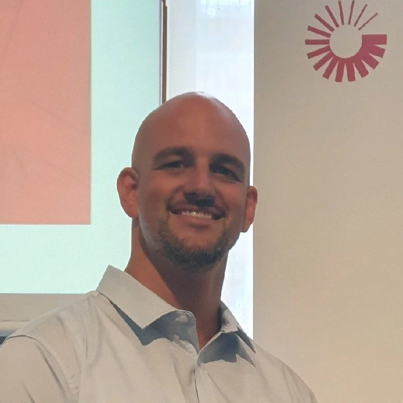 Man with a bald head and goatee smiling in a light-colored dress shirt, standing in front of a projection screen and a wall with a red circular design.