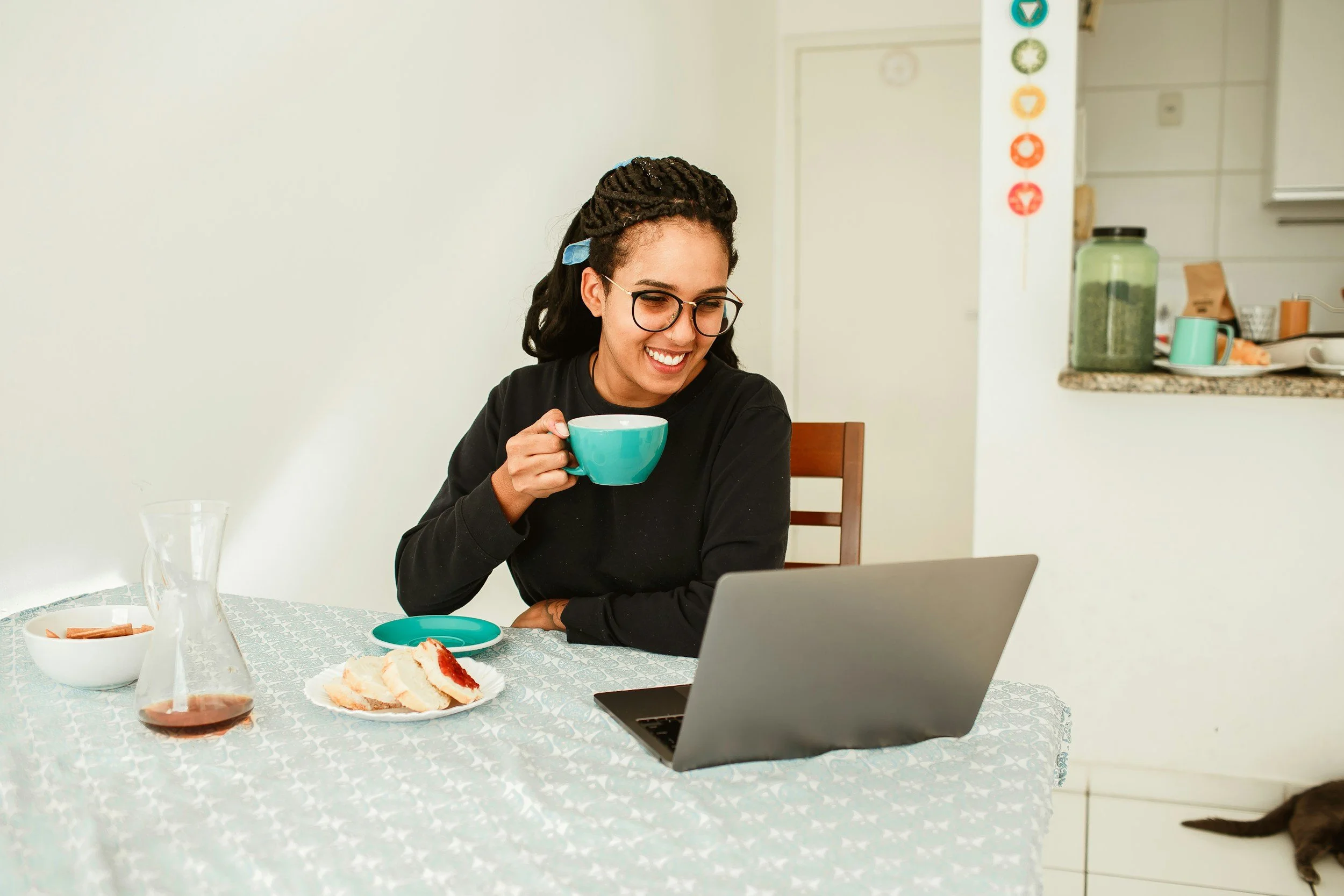 A woman with glasses and dreadlocks sitting at a table, smiling, while holding a teal mug, with a plate of cake, a bowl, and a glass pitcher on the table, looking at a laptop.