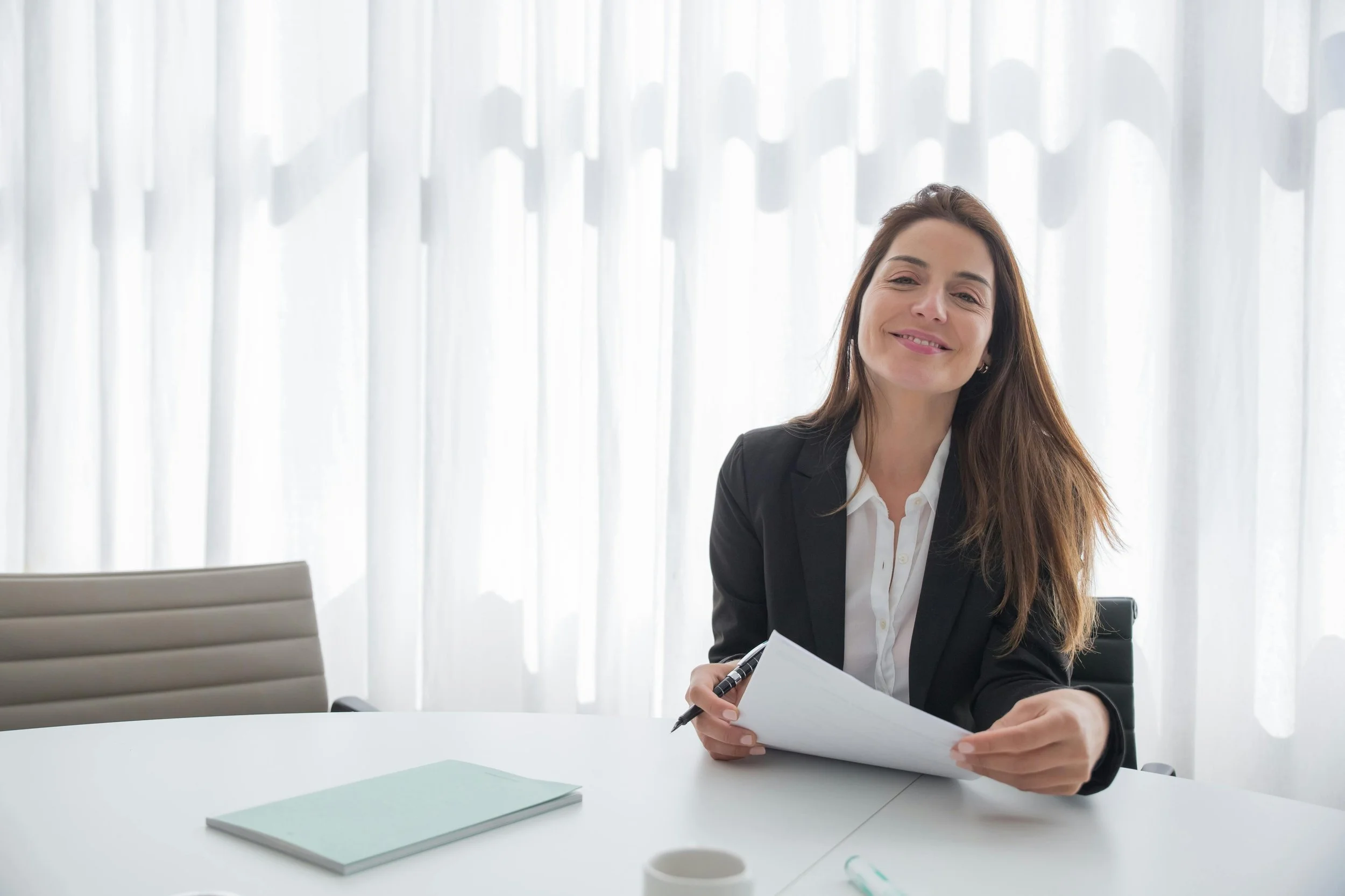 Businesswoman sitting at a white table, smiling, holding documents with sunlight filtering through curtains in the background.