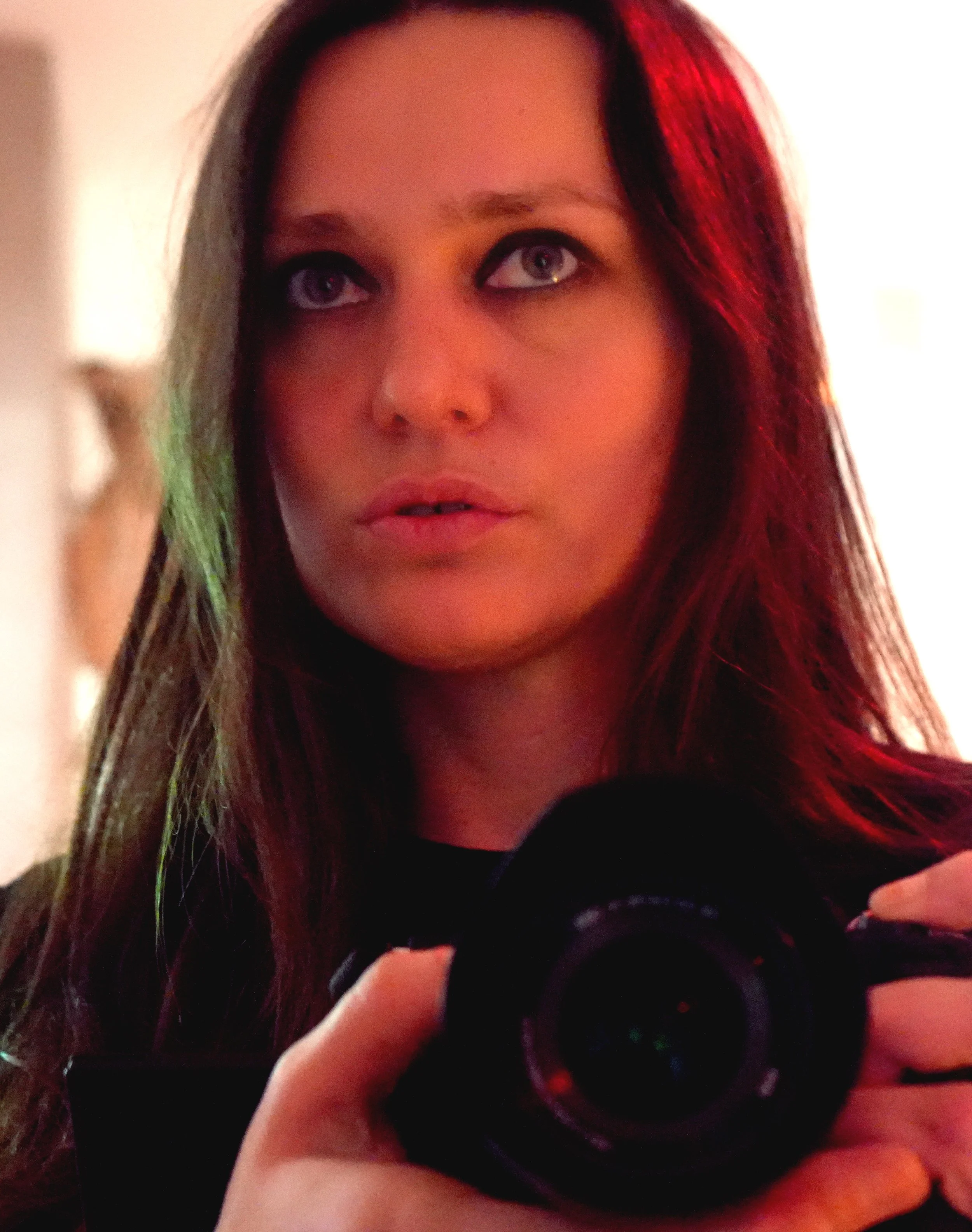 A woman with long brown hair holding a camera in front of a mirror, taking a selfie.
