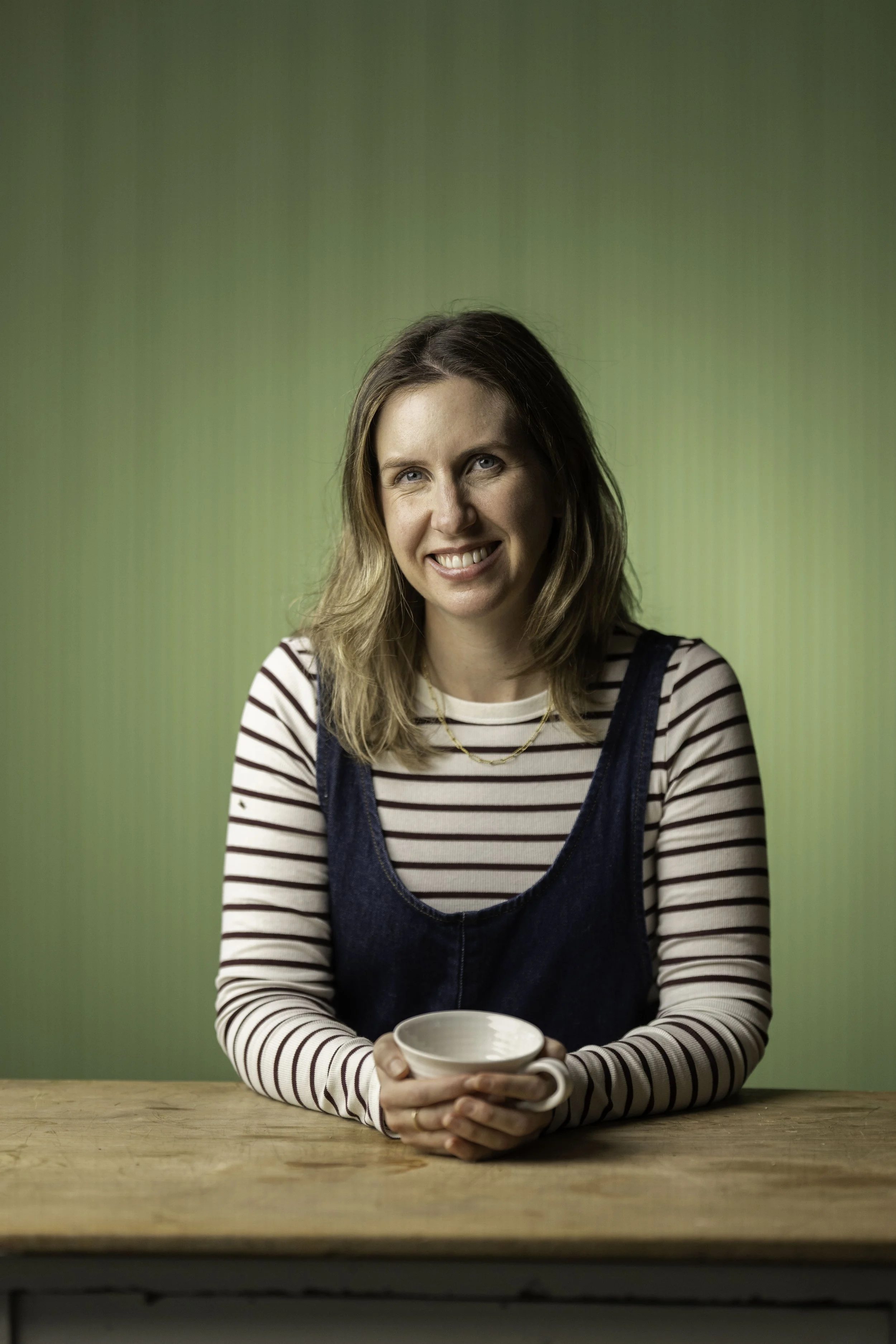 A young woman with shoulder-length blonde hair, wearing a striped long-sleeve shirt and denim apron, sitting at a wooden table with a white mug in her hands, smiling at the camera against a green background.