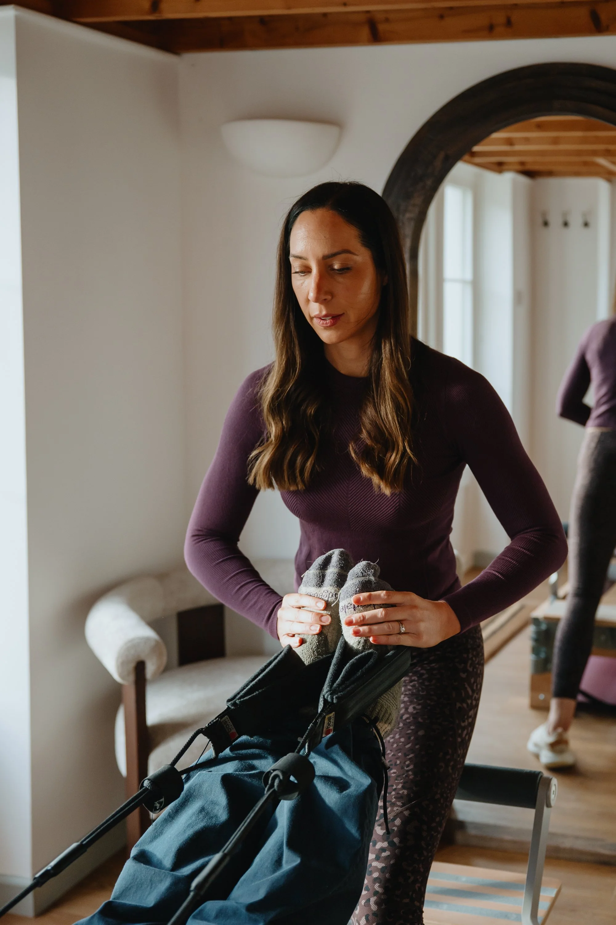 A woman with long dark hair, wearing a purple long-sleeve shirt and patterned pants, stands indoors with a stroller, holding a pair of gray socks and looking down.