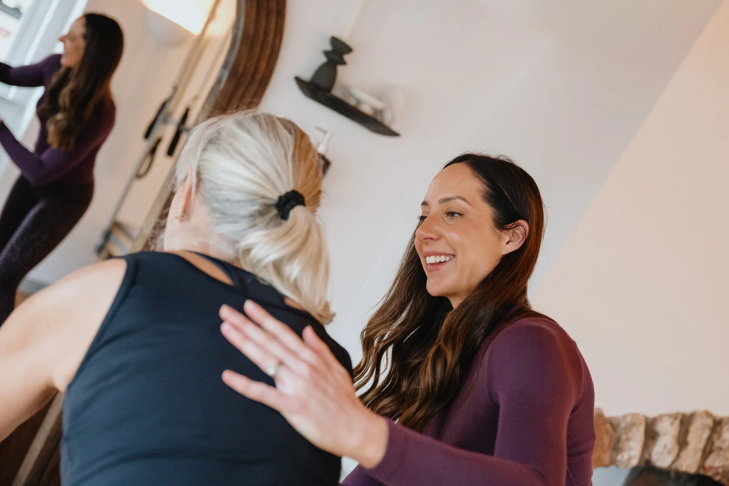 Two women smiling and embracing in a cozy indoor setting, with a third woman in the background near a doorway