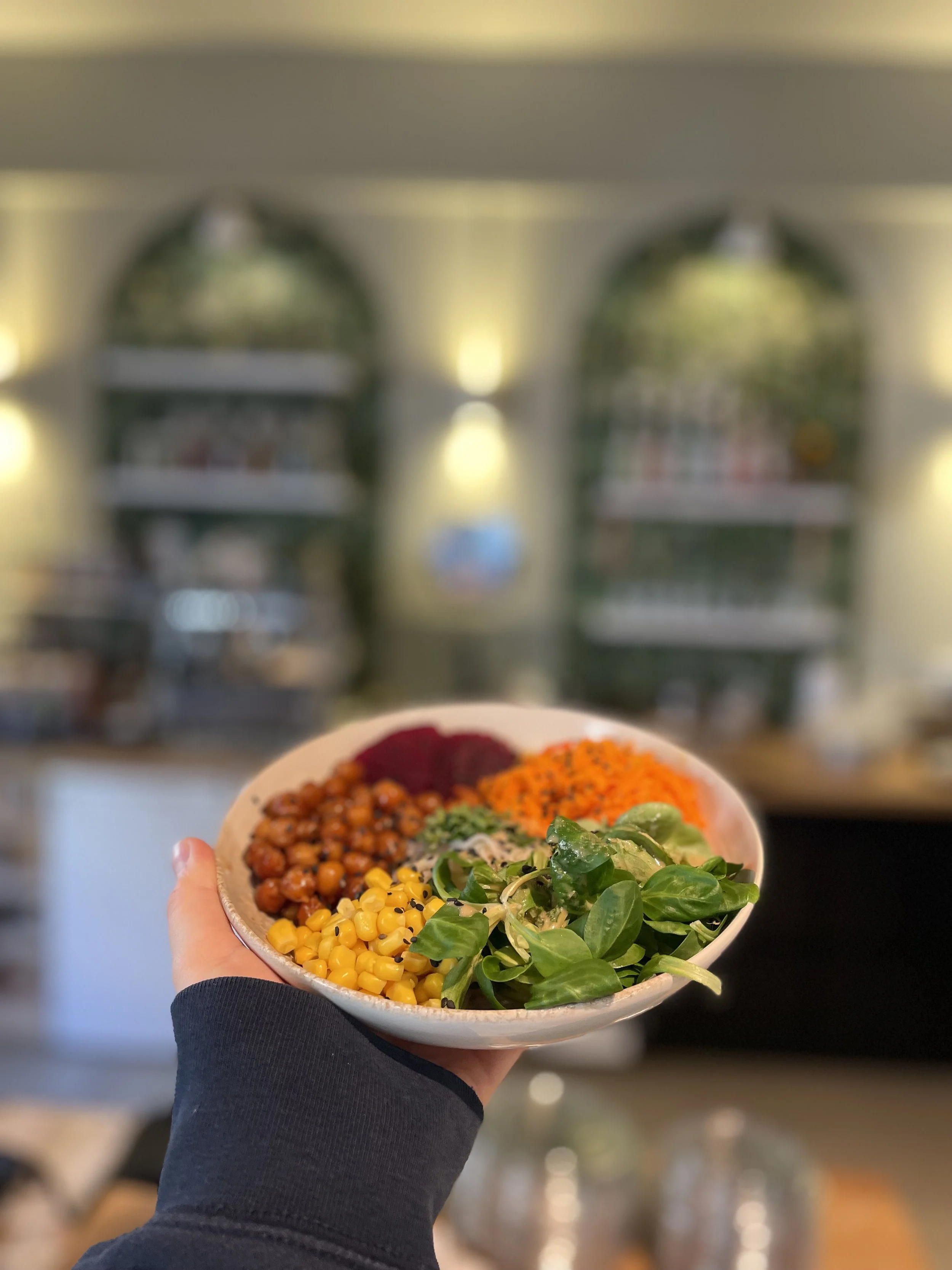 Hand hält eine Schüssel mit buntem Salat, dazu Mais, Kichererbsen, Rote Beete, Blattgemüse und Karotten. Im Hintergrund unscharfer Raum mit Regal und Beleuchtung.