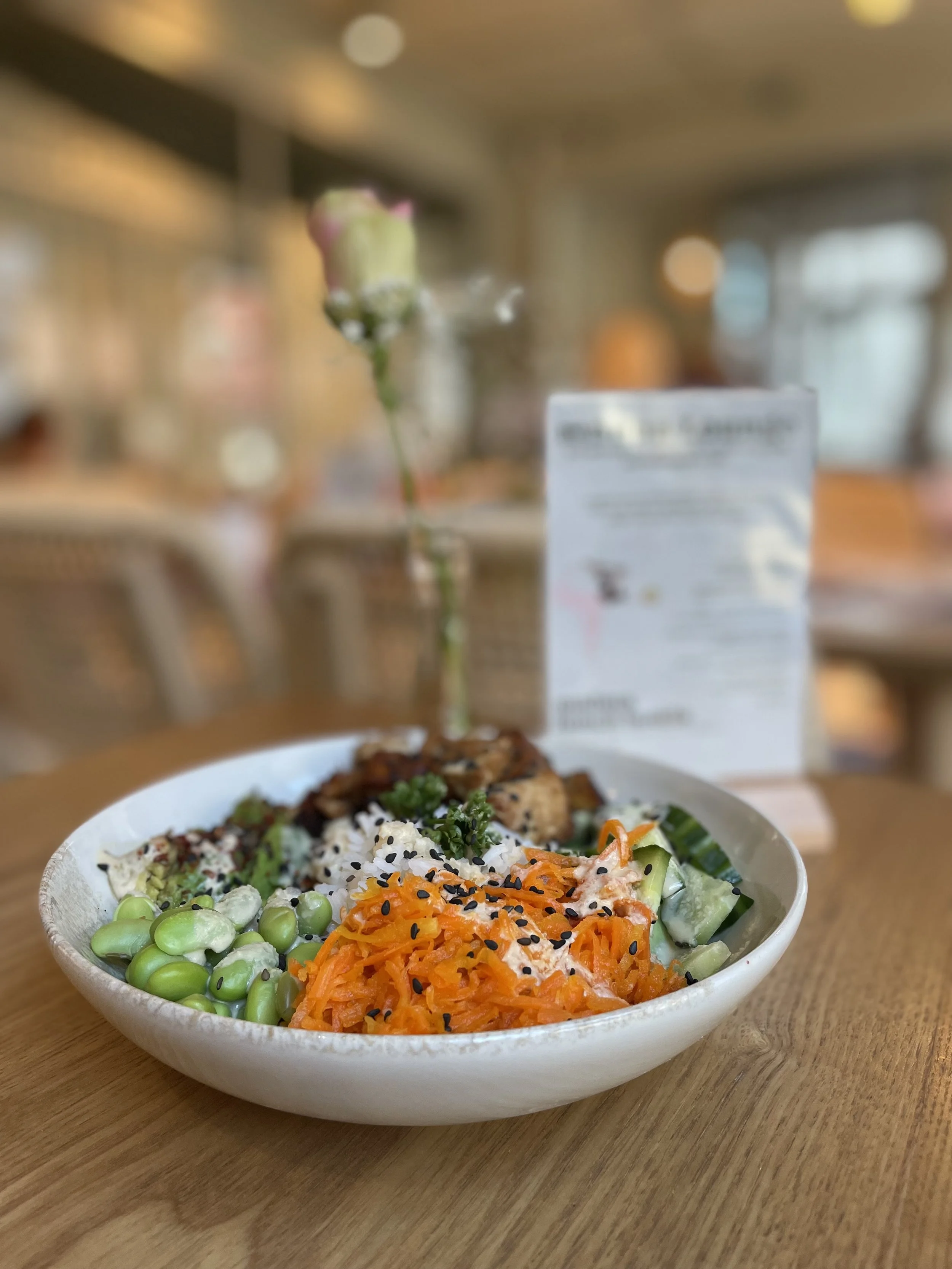 Buddha-Bowl mit Reis, geraspelten Karotten, Gurken, Edamame und Hähnchen, mit einer Blume in einer Vase im Hintergrund