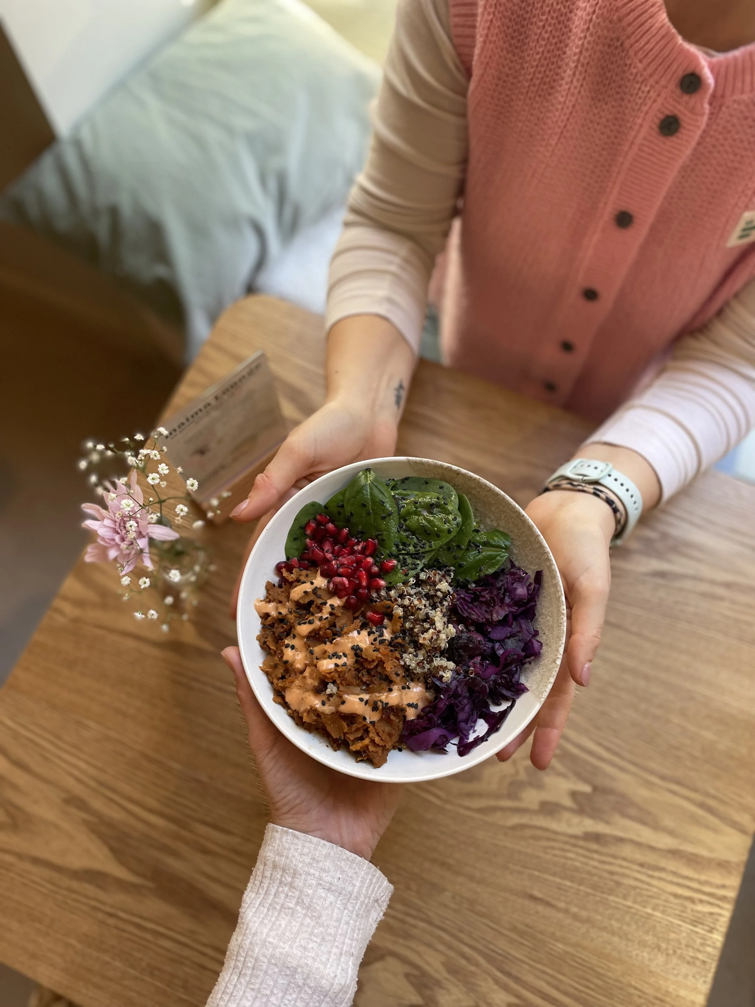 Person hält eine Bowl mit buntem Salat mit Spinat, roten Granatapürekernen, getrockneten Tomaten, Quinoa und Rotkohl, während eine andere Person an einem Holztisch sitzt.