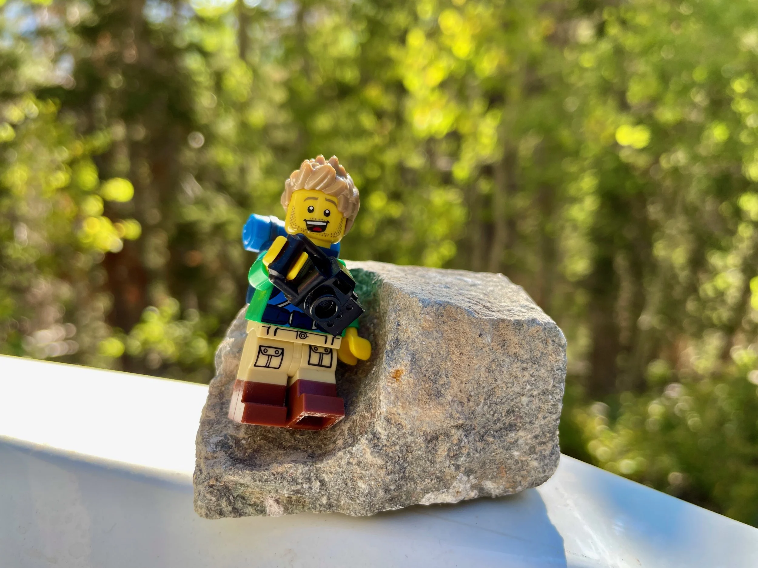 LEGO photographer minifigure with a camera and backpack sitting on a rock outdoors, surrounded by sunlit green trees.