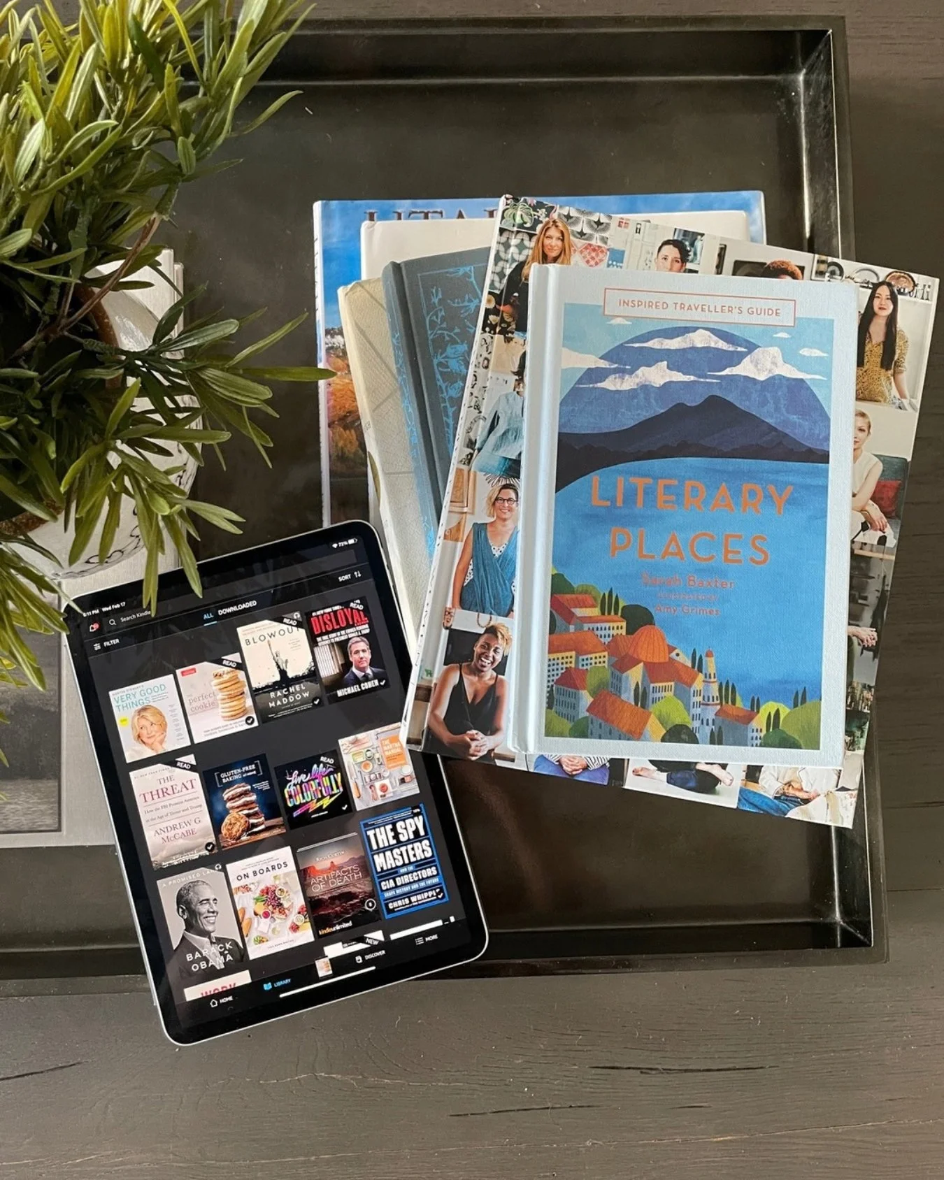 Stack of books about travel and other interests beside an iPad displaying a digital library of books.
