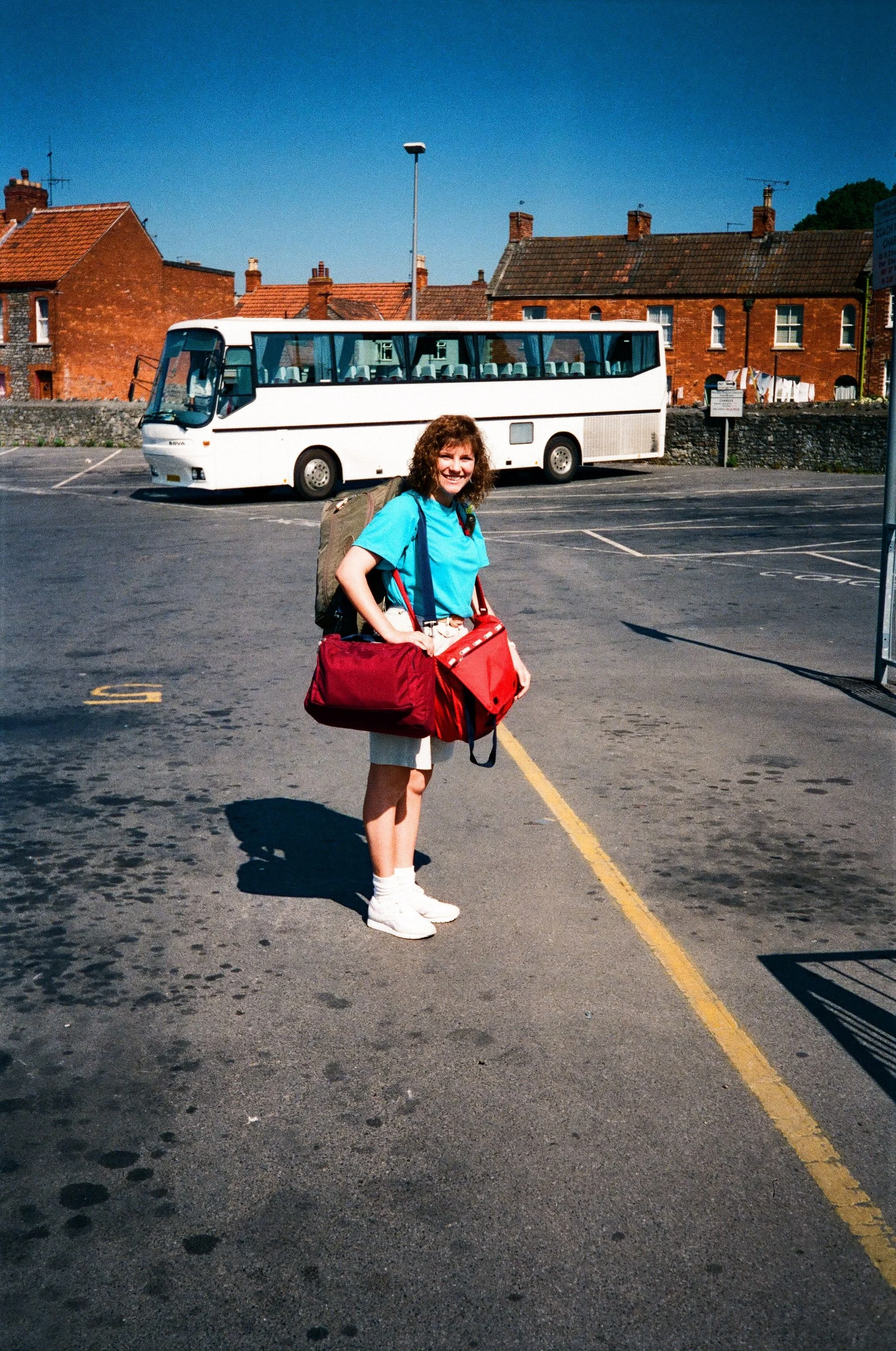 College student wearing a turquoise shirt and carrying travel bags in a parking lot in England during a 1989 study abroad program, with a white tour bus and brick buildings in the background.