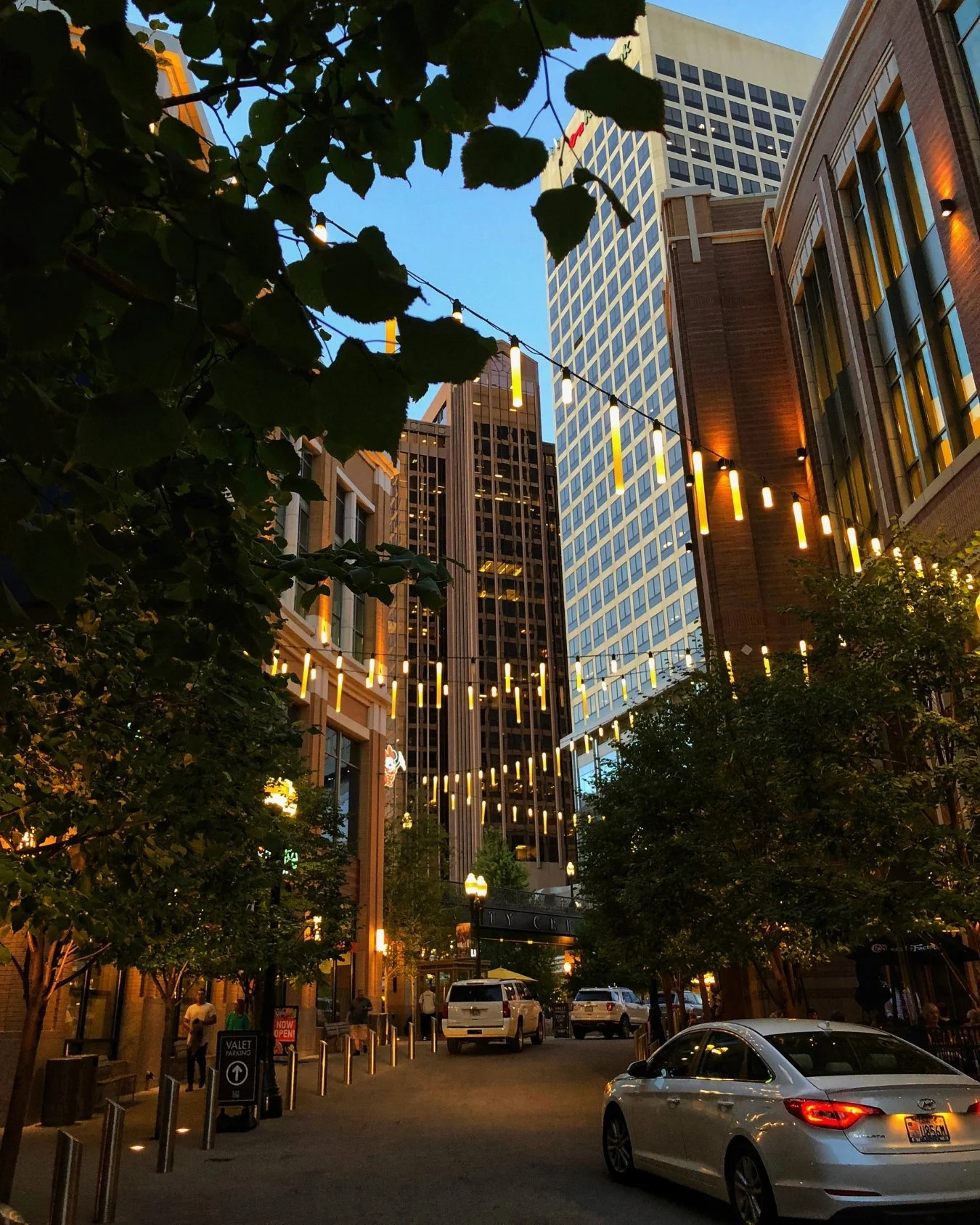 Evening view of downtown Salt Lake City, Utah, with glowing string lights suspended between buildings, tree-lined streets, and modern skyscrapers creating a warm, inviting city atmosphere.