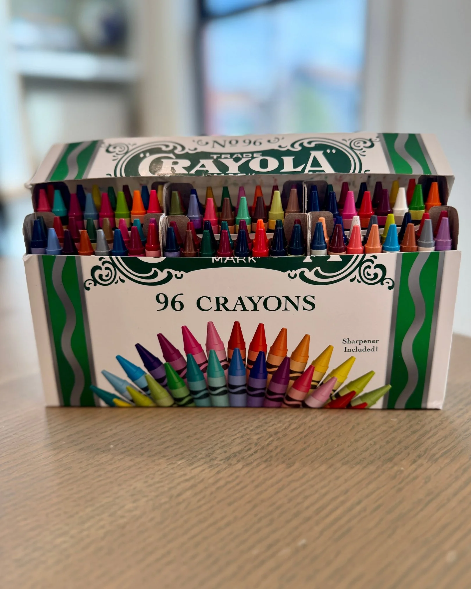 Open box of 96 Crayola crayons displayed on a wooden table with colorful crayons arranged inside.