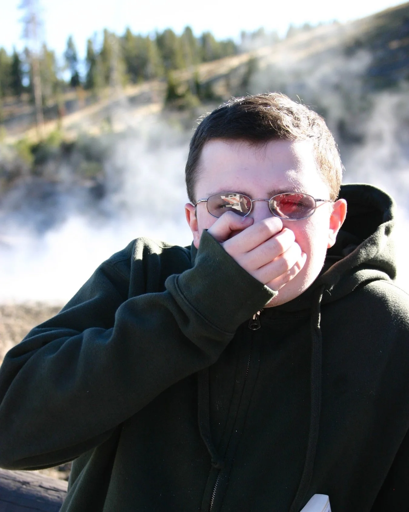 Person reacting to the strong sulfur smell at Dragon Mouth Spring in Yellowstone National Park, with rising steam from the geothermal spring in the background.