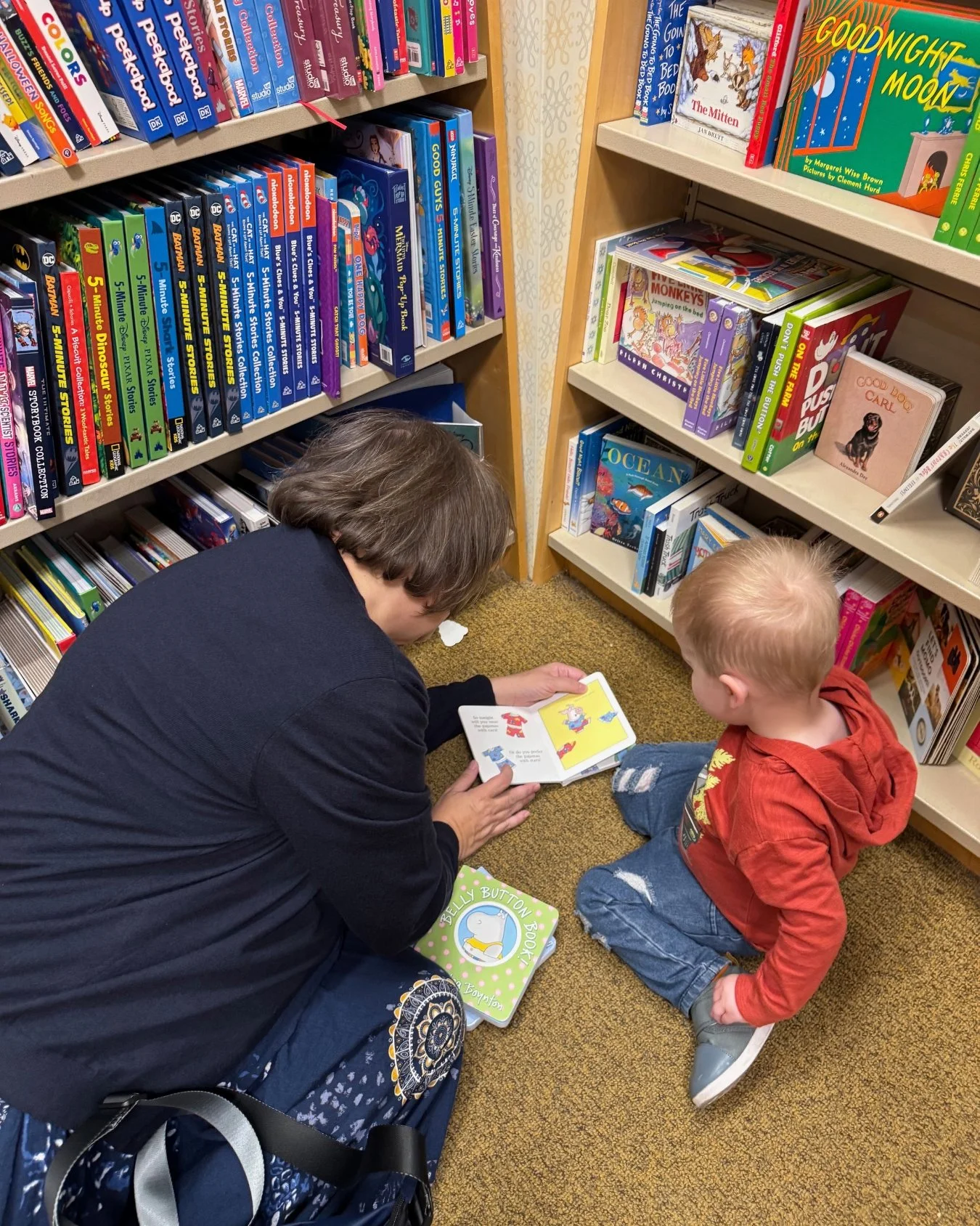 Grandmother and young grandson sitting together between bookstore shelves and looking at a children's book.