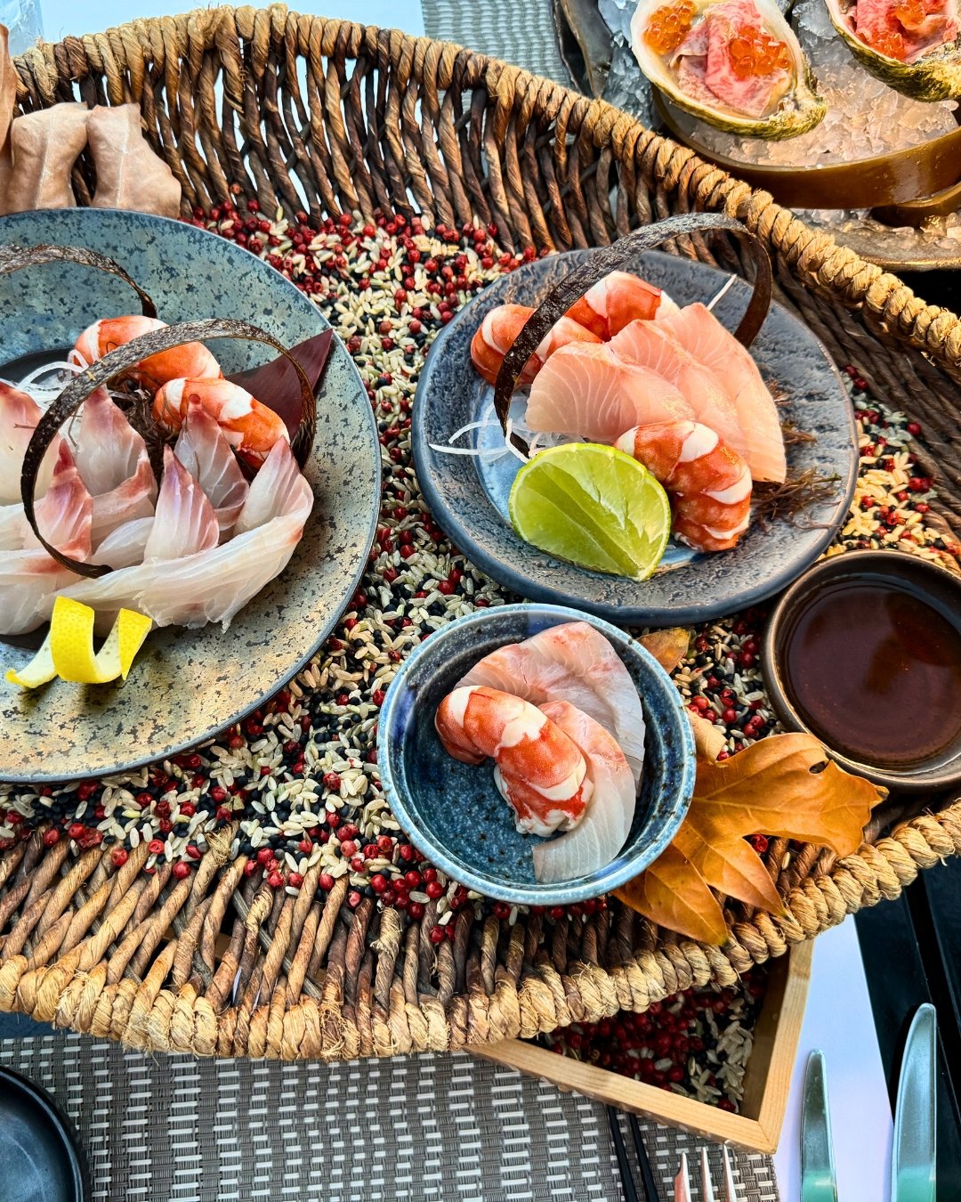 Assorted sashimi and seafood arranged in ceramic bowls on a woven tray filled with colorful grains, garnished with citrus slices and autumn leaves