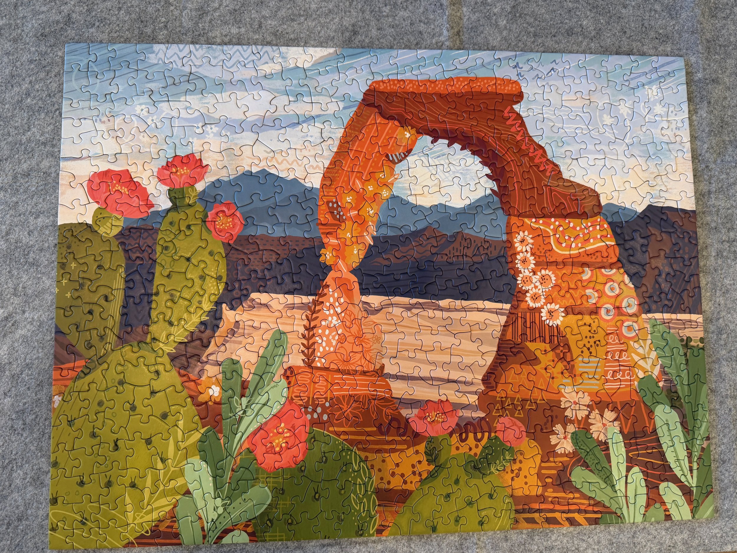 Completed jigsaw puzzle featuring a colorful illustrated view of Delicate Arch surrounded by desert plants and mountains.