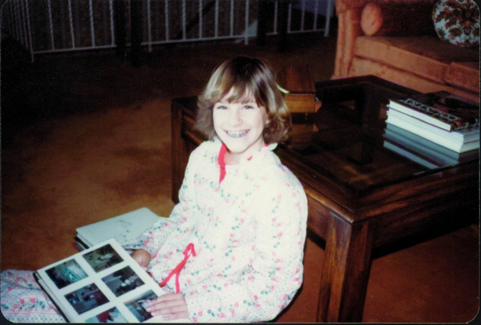 Smiling young girl wearing floral pajamas and braces, sitting on the floor and looking through a family photo album beside a wooden coffee table.