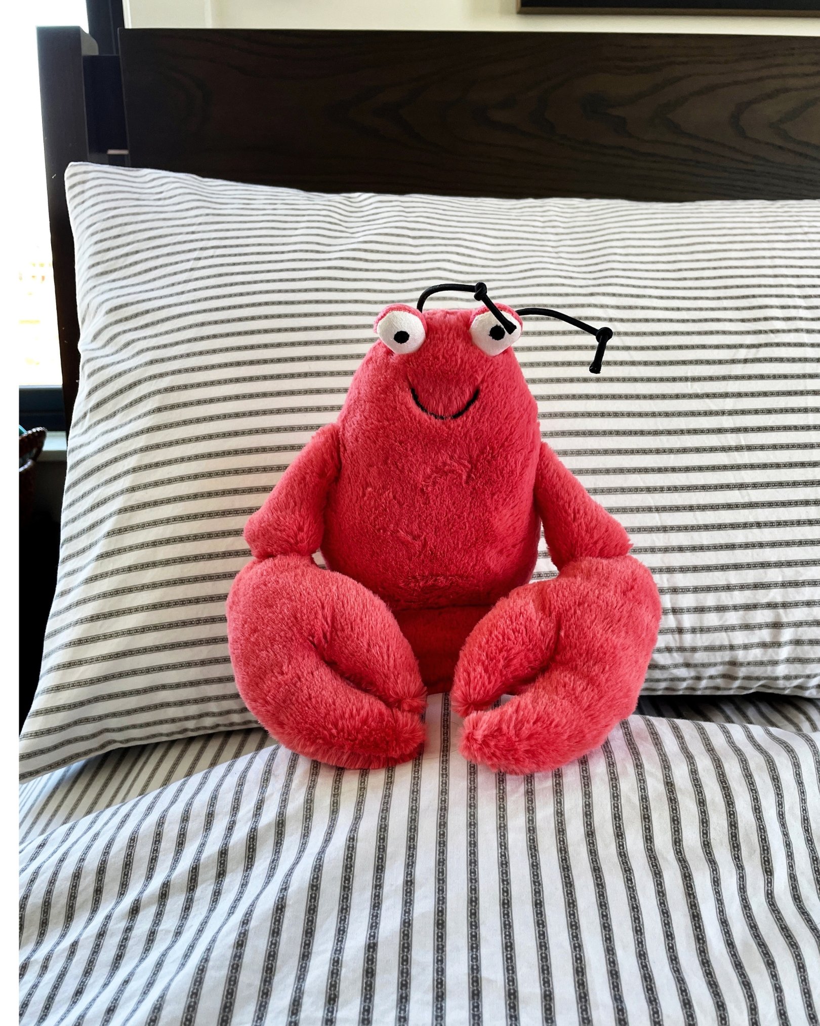 Red lobster stuffed animal sitting on a striped bed with a smiling face and playful expression, ready for a cozy bedtime adventure.