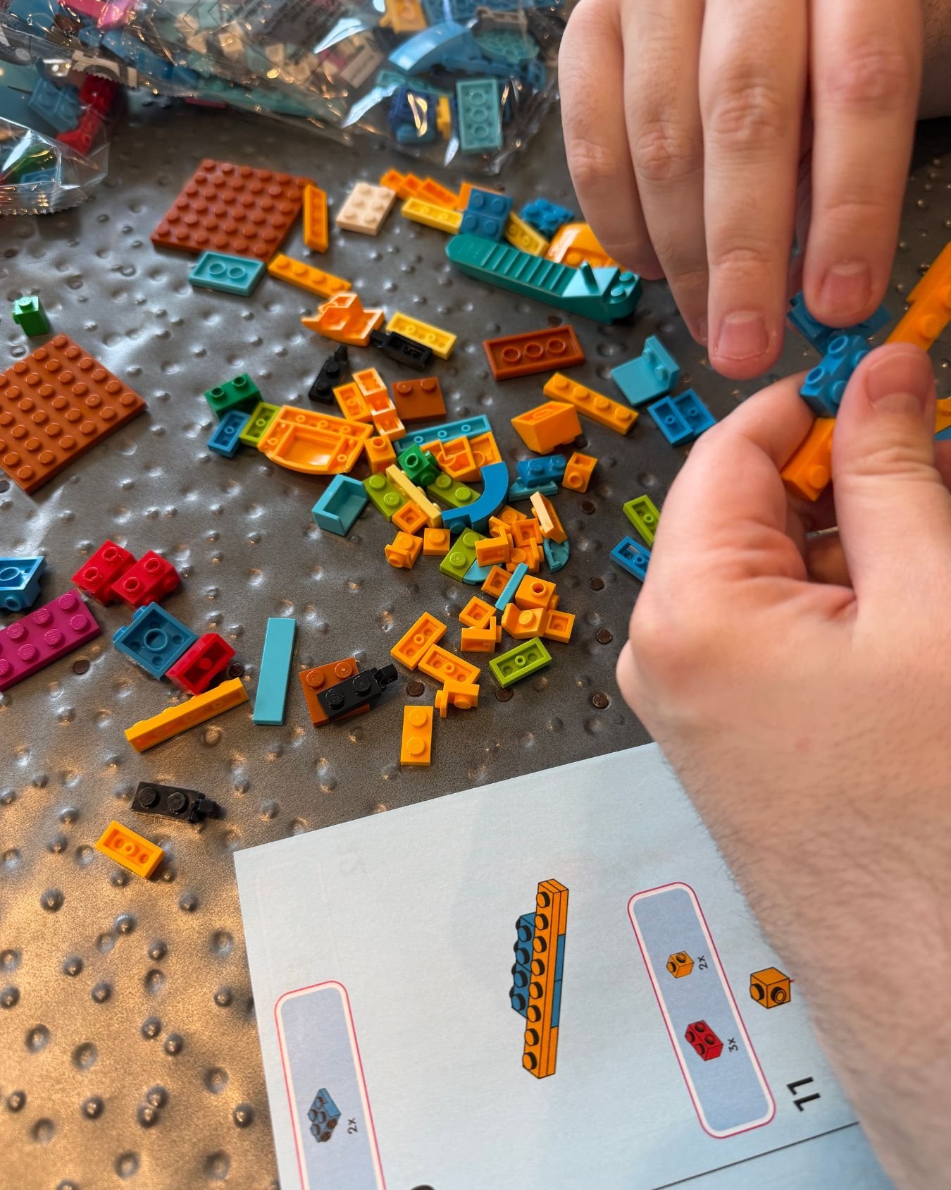 Hands building a colorful LEGO set together with loose bricks and an instruction booklet spread across the table.