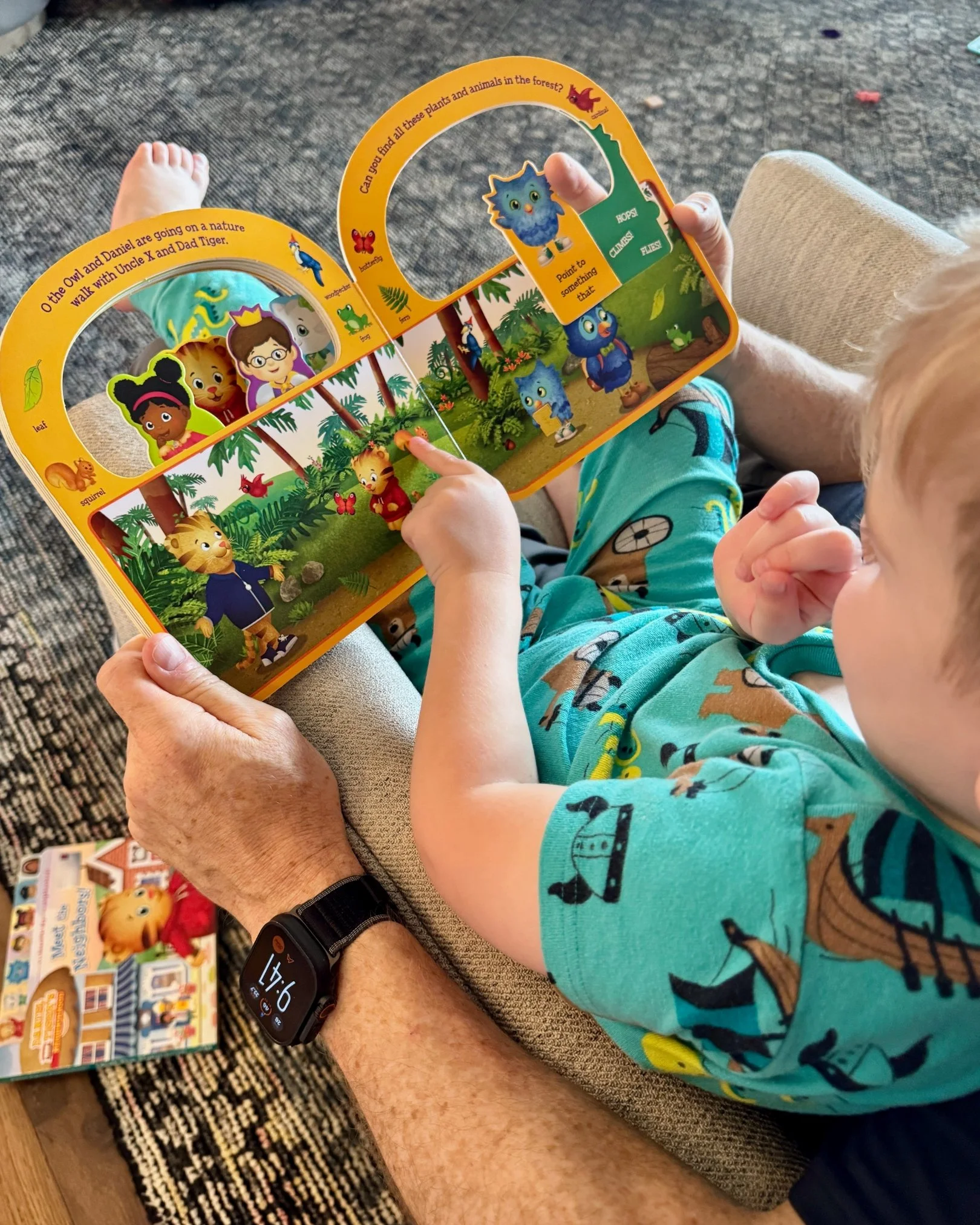 Young child sitting on an adult's lap while reading an interactive Daniel Tiger board book together during quiet playtime.