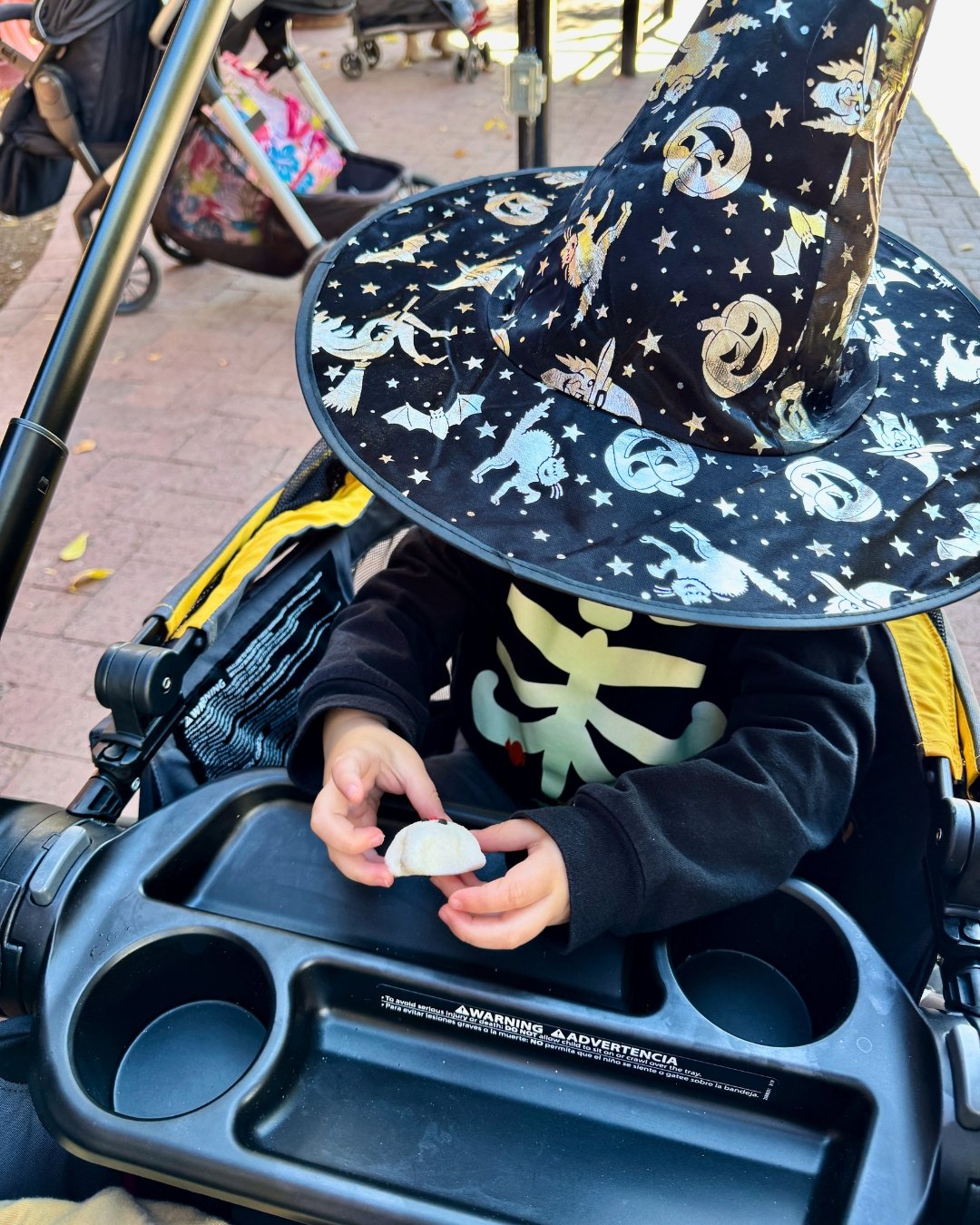 Young child wearing an oversized Halloween witch hat and skeleton sweatshirt while examining a small white object in a stroller.