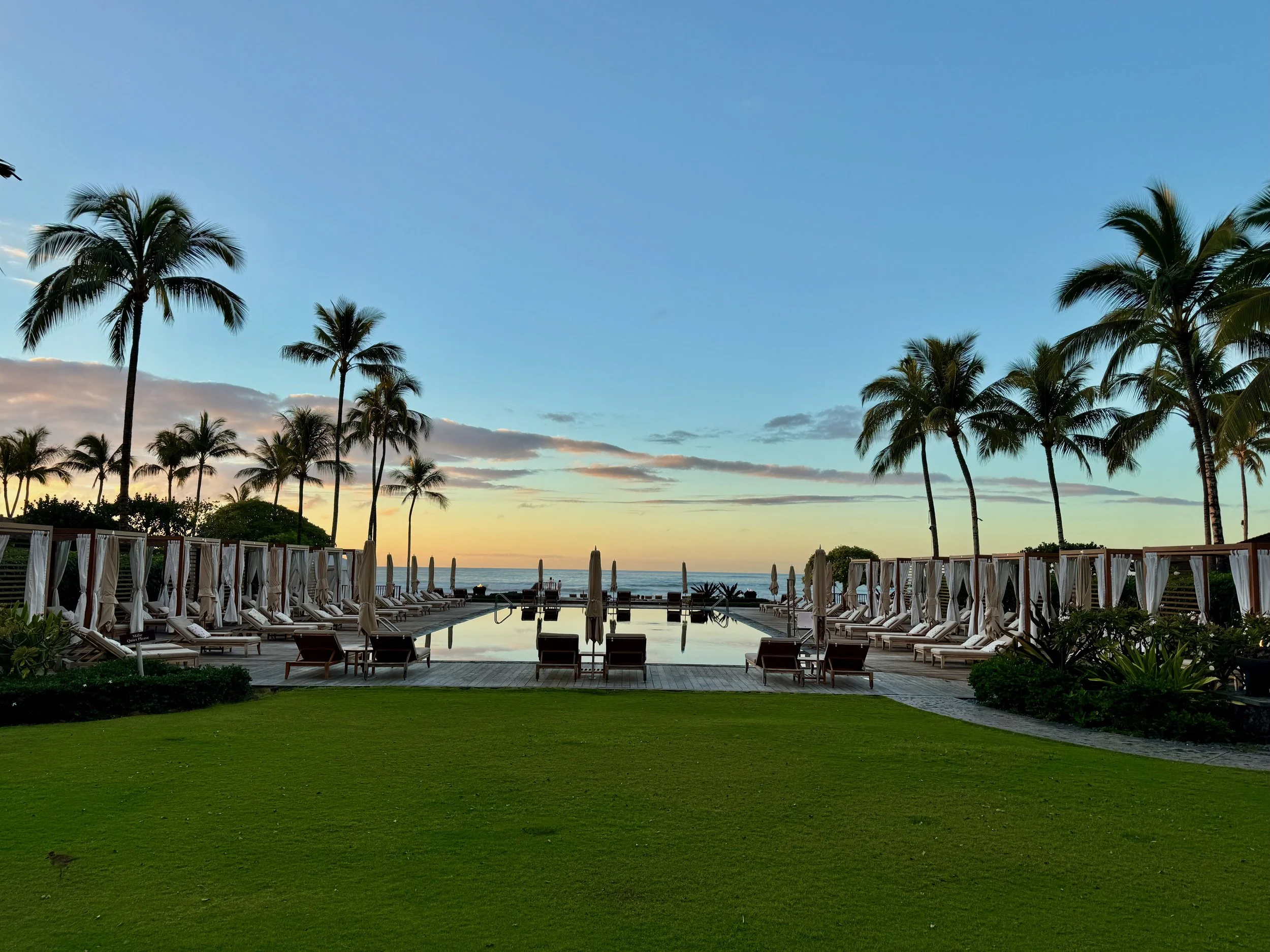 Resort pool surrounded by palm trees overlooking the ocean at sunrise in Hawaii.