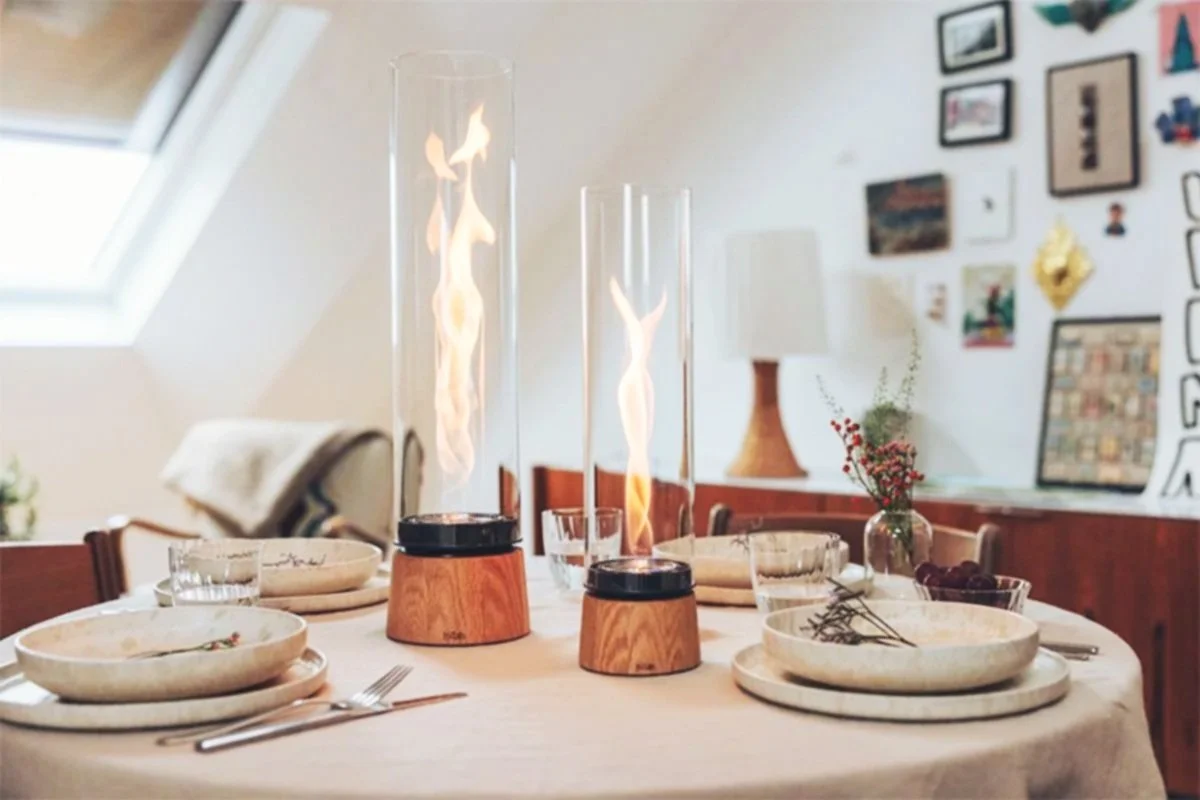 Une table à manger décorée avec deux lanternes à flamme vacillante, vaisselle en céramique, et un décor mural avec plusieurs photos et œuvres d'art dans une pièce éclairée par une fenêtre de toit.
