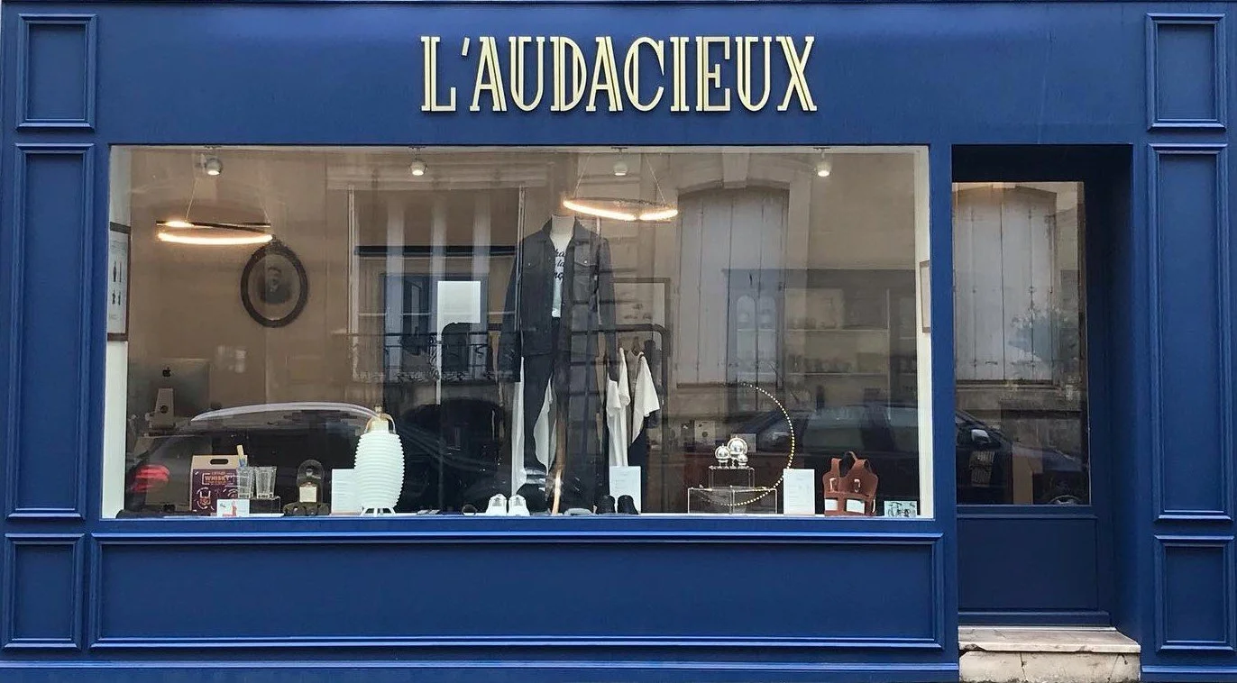 Vitrine d'un magasin nommé 'L'AUDACIEUX' avec des vêtements et des accessoires exposés