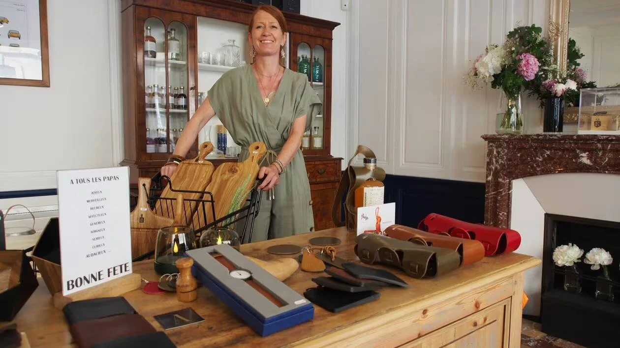 Femme souriante derrière un comptoir en bois avec des articles en cuir, un vase de fleurs colorées sur la droite, un panneau avec une liste de noms et une phrase 'Bonne Fête' sur la gauche, dans un intérieur élégant avec un grand meuble vitré et une cheminée en marbre.