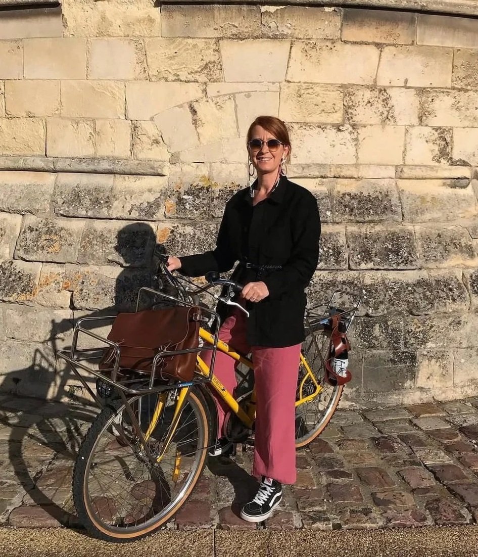 Femme souriante avec lunettes de soleil, vêtements noirs et pantalon rose, posant avec un vélo jaune équipée d'un panier en métal, devant un mur en pierre.