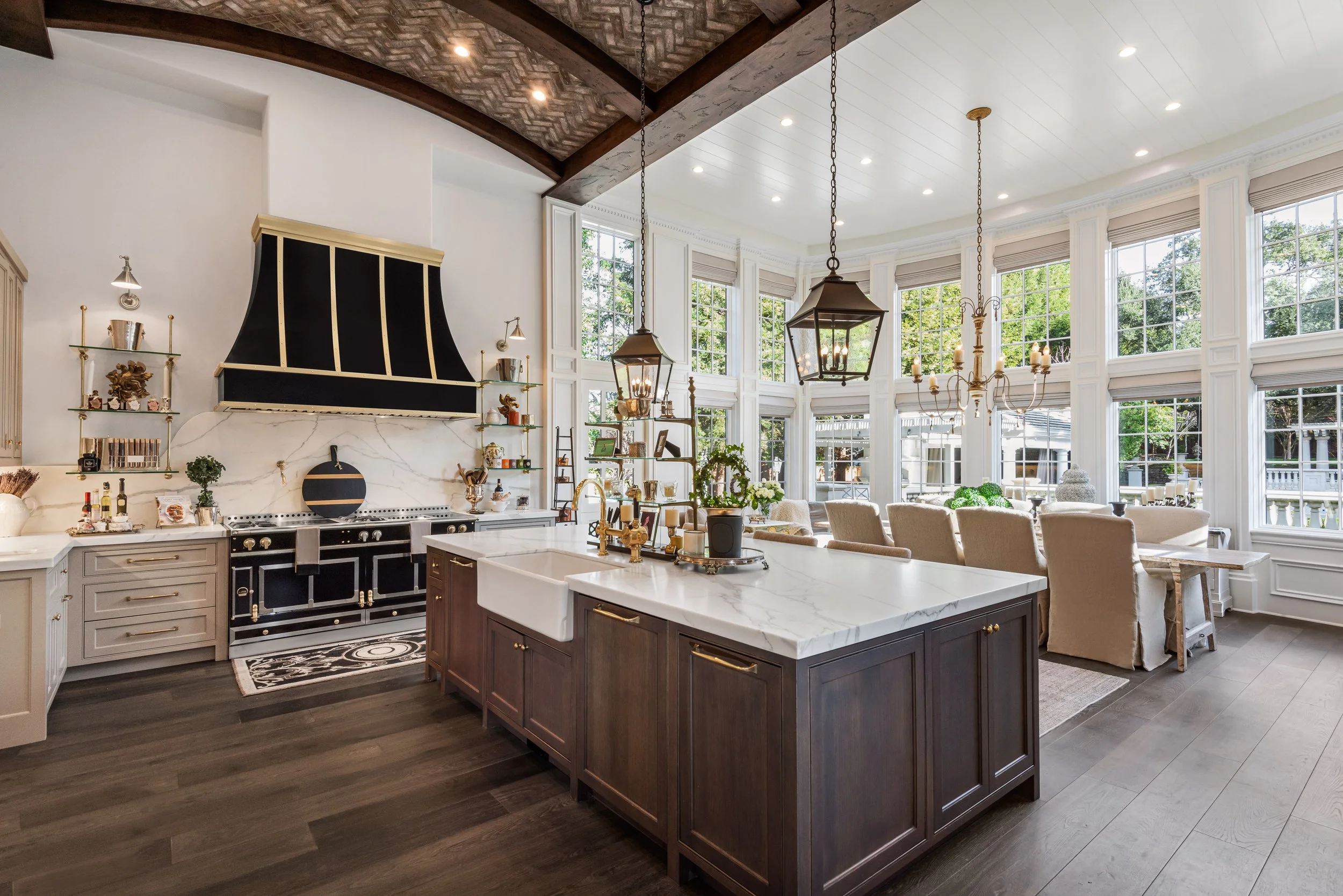 A spacious, bright kitchen with a large center island with a marble top, a black and white stove with gold accents, and a vaulted ceiling with wooden beams. The open dining area has a white sectional sofa and large windows showing greenery outside.