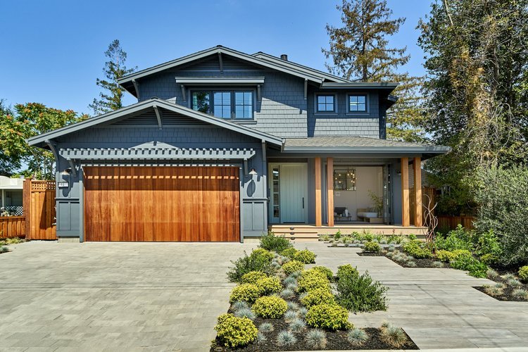 Modern two-story house with gray siding, a wooden garage door, and a landscaped front yard with shrubs and a paved driveway.