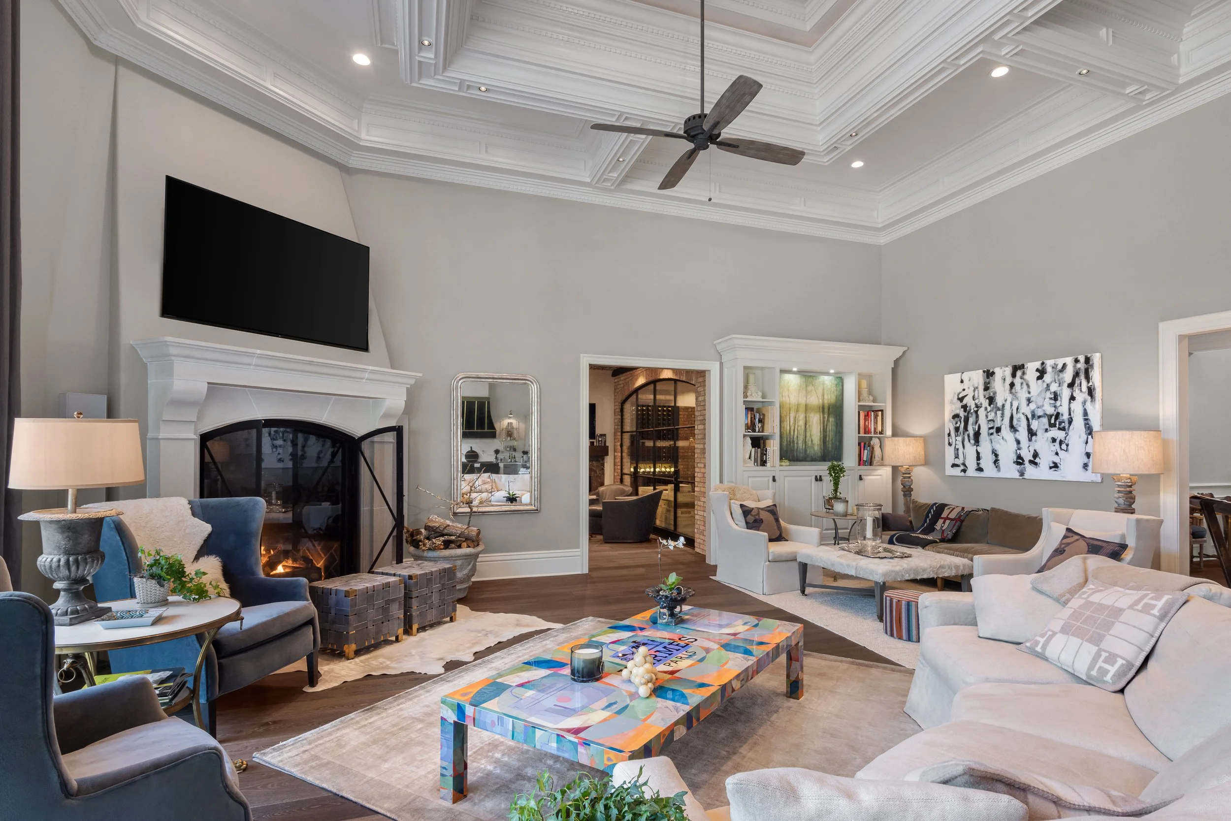 Living room with a high ceiling, ceiling fan, large TV above fireplace, fireplace with logs, multiple sofas and armchairs, colorful coffee table, artwork, and lamps.