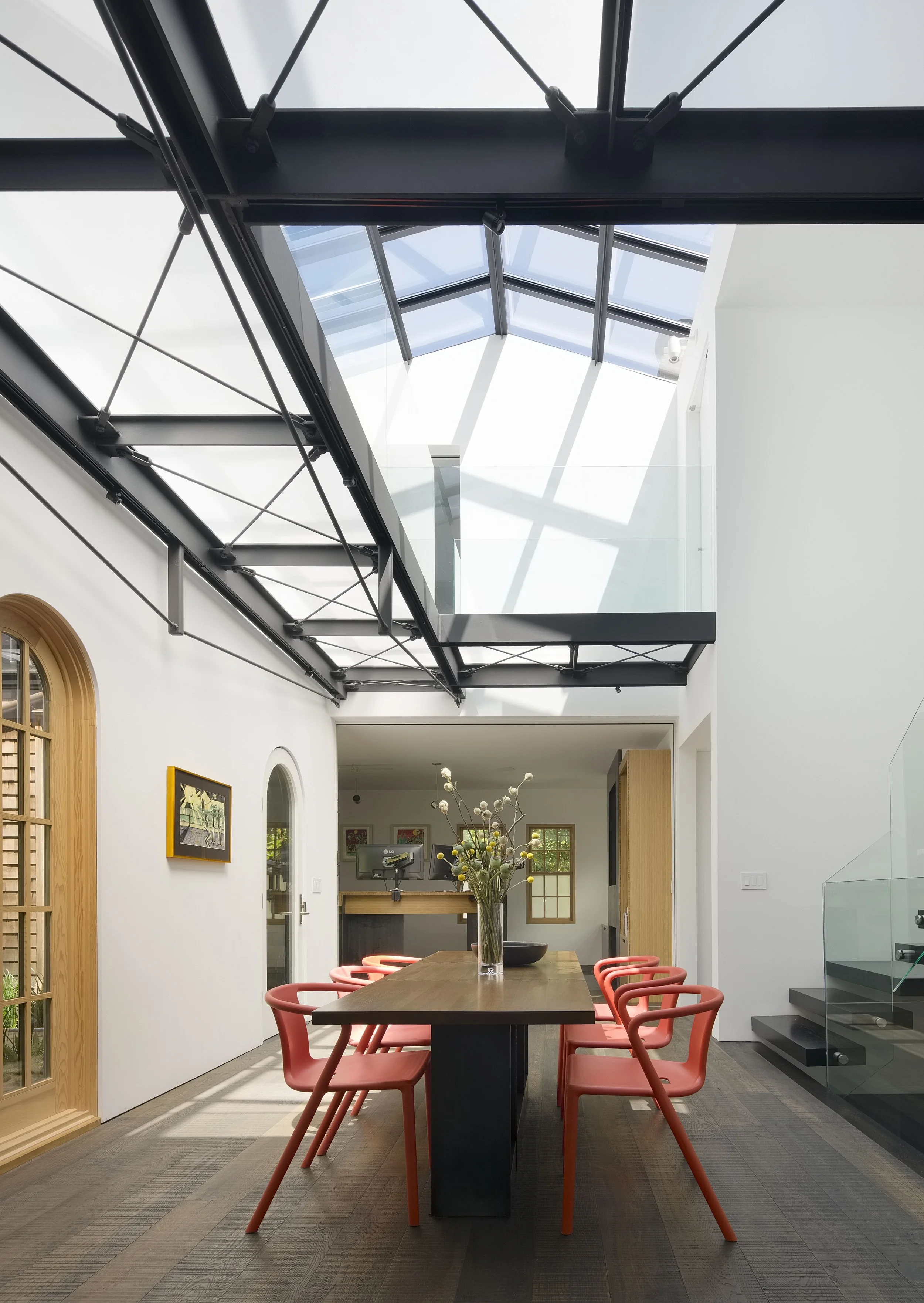 Modern dining room with a wooden table, red chairs, a glass vase with flowers, white walls, and a glass ceiling allowing natural light.