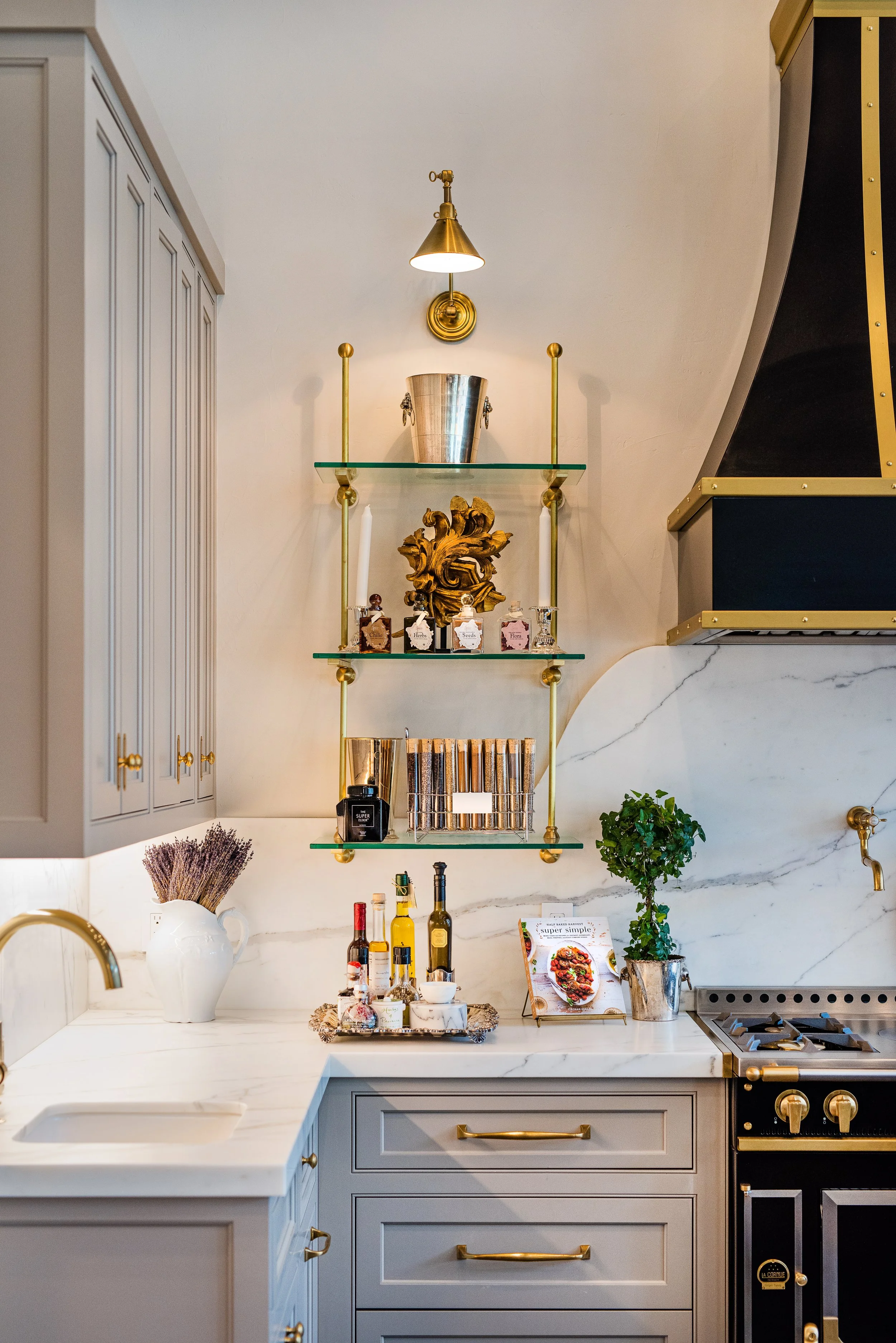 Kitchen with grey cabinets, white marble countertops, black and gold stove, and decorative open shelves holding vases, books, and small jars; with a potted plant and cookbook on the counter.