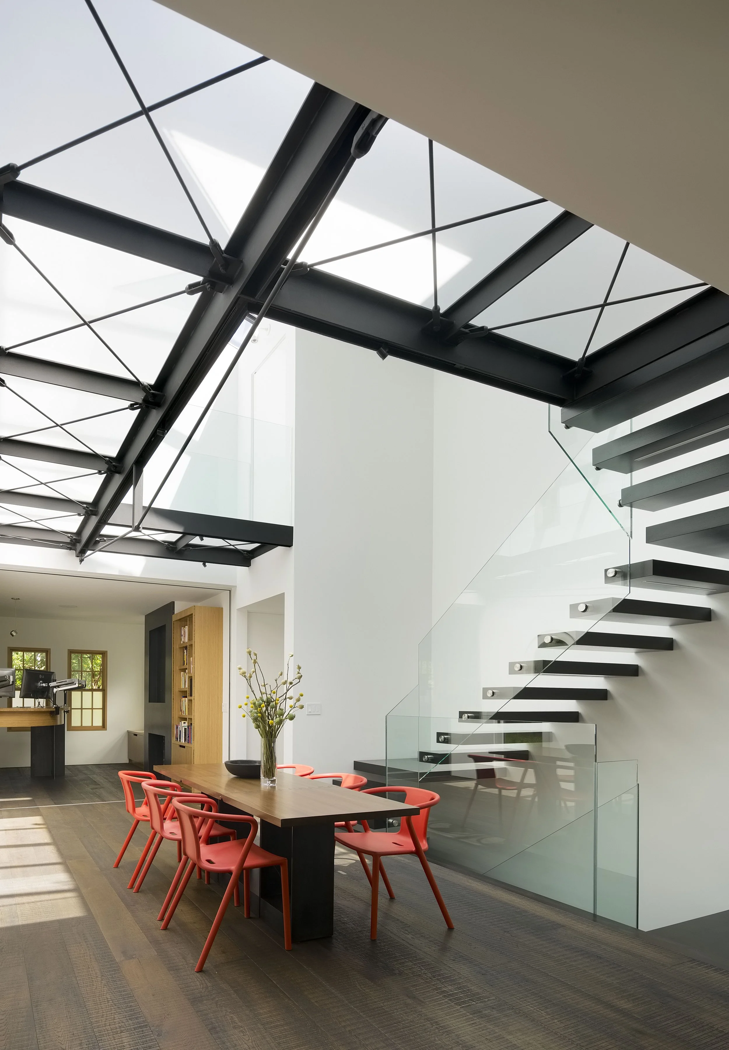 Modern dining room with dark wood table, coral chairs, glass staircase, skylight ceiling, and a corner with bookshelves.