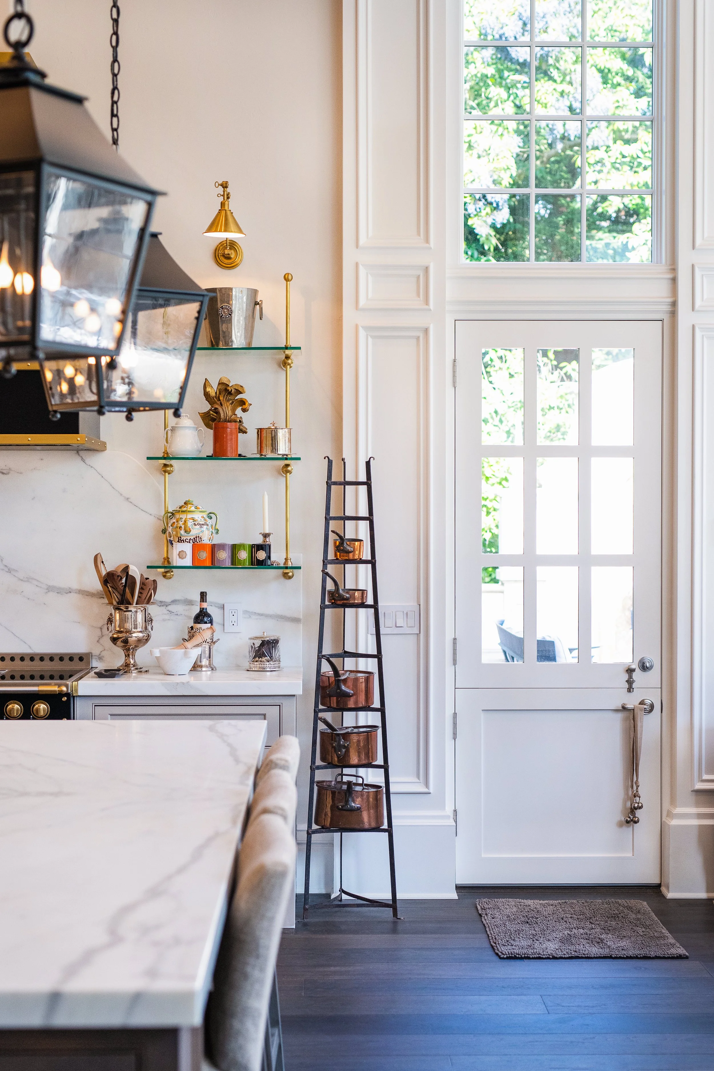 Kitchen door with large window above, white cabinetry, and a black ladder shelf holding small copper pots, decorative jars, and a small sculpture, with a marble countertop and dark wood flooring.
