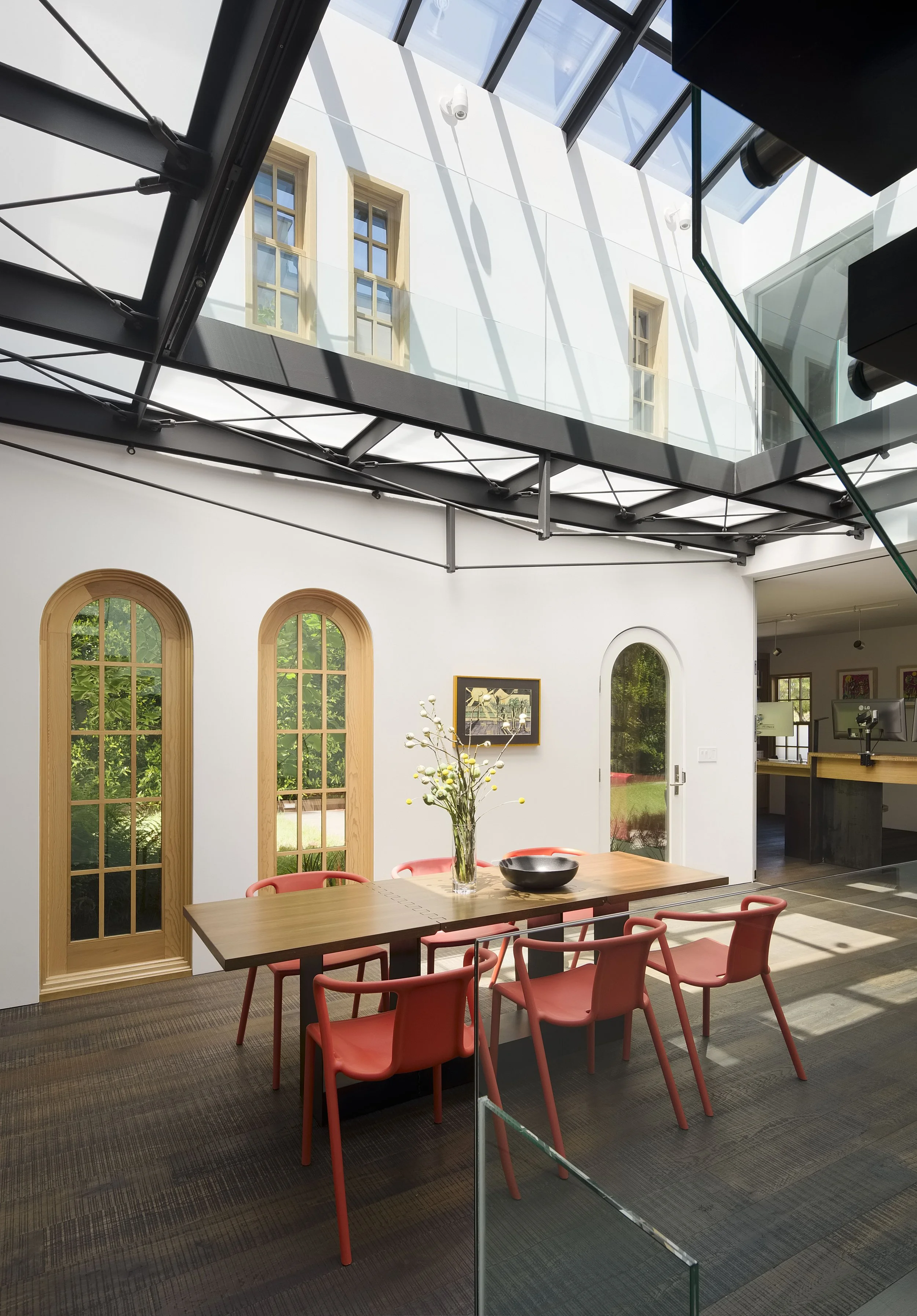 Modern dining room with a wooden table surrounded by coral-colored chairs, a floral centerpiece, and a white wall with arched windows, natural light, and a staircase leading to an upper level.
