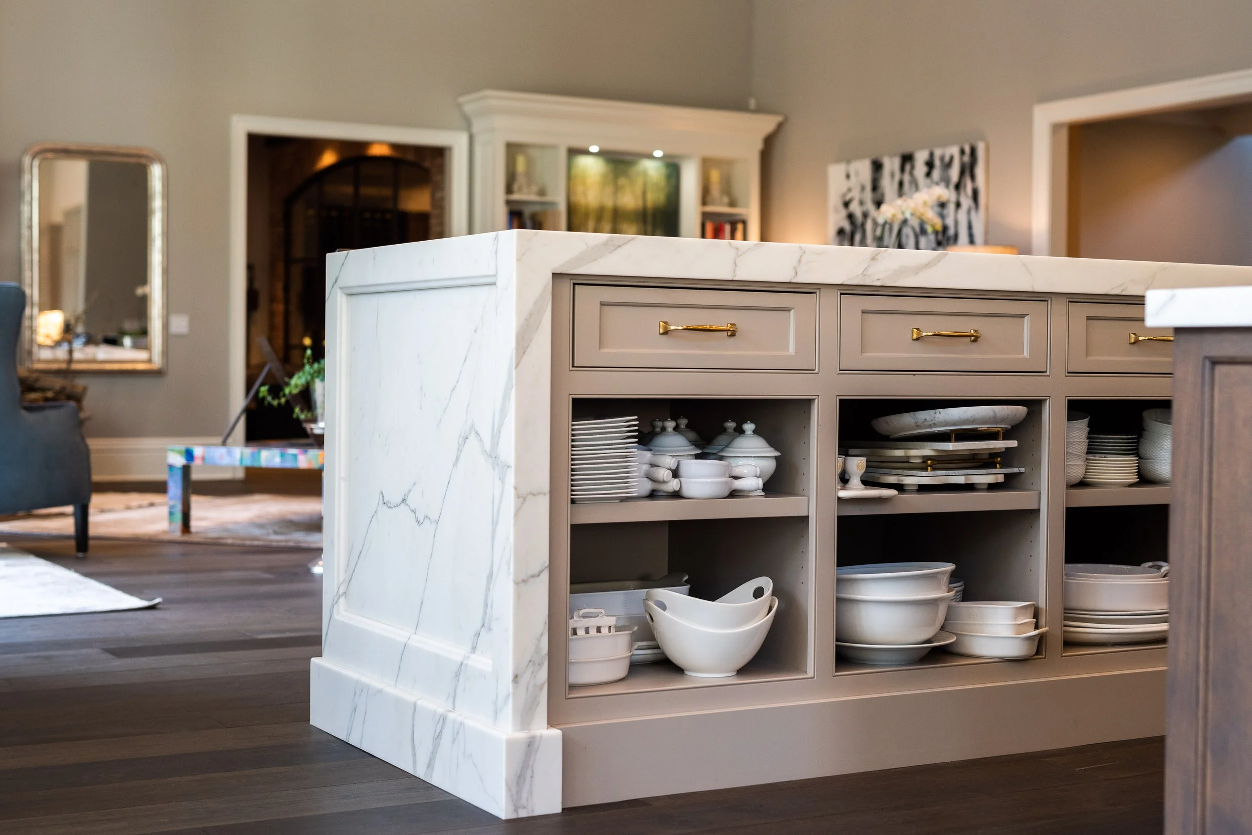 Kitchen island with beige cabinets and white marble top, containing stacked white dishes and bowls, in a modern living space.