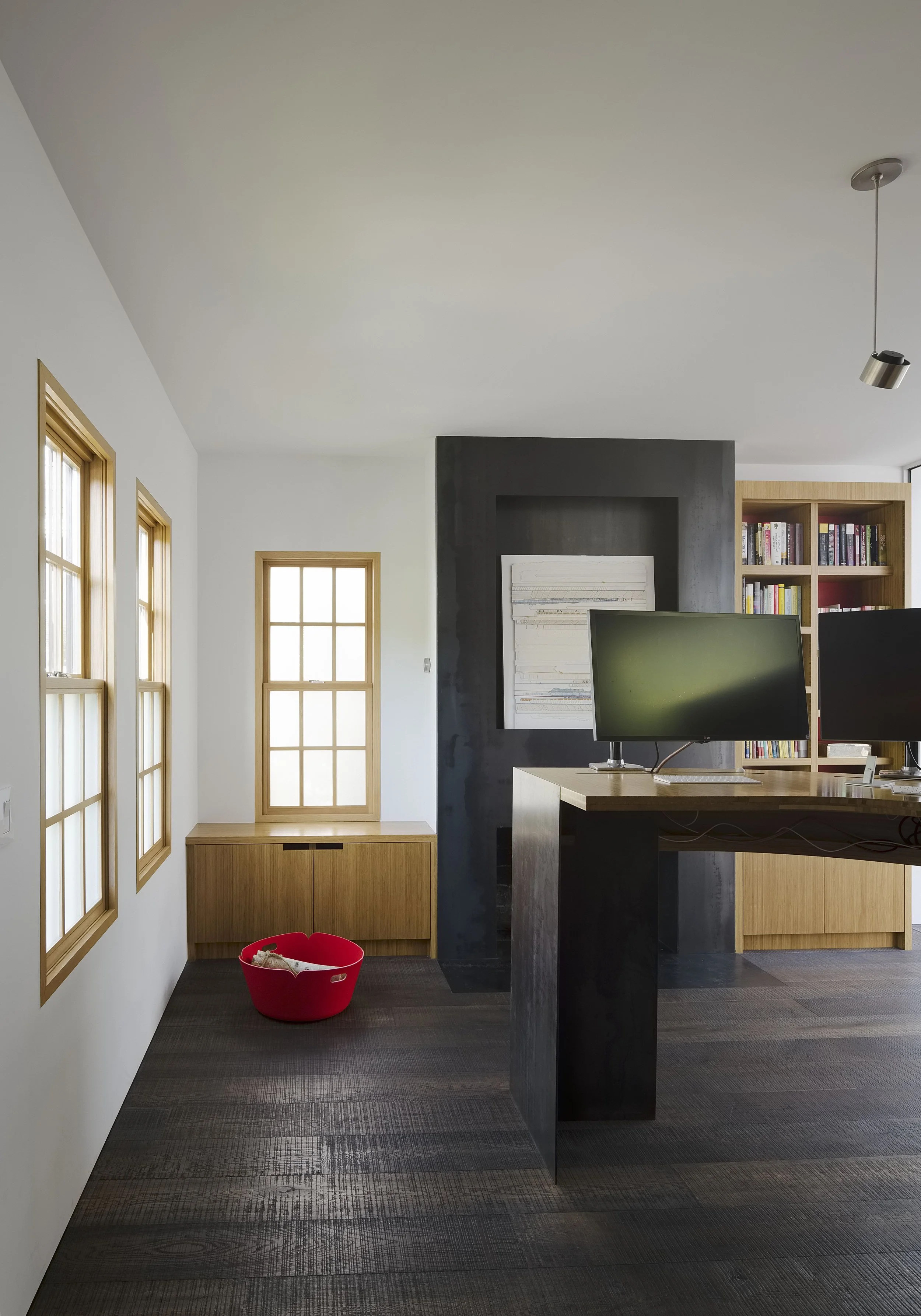 Modern home office with three windows, a red pet bed with a dog inside, a black accent wall, wooden cabinetry, and two computer monitors on a wooden desk.